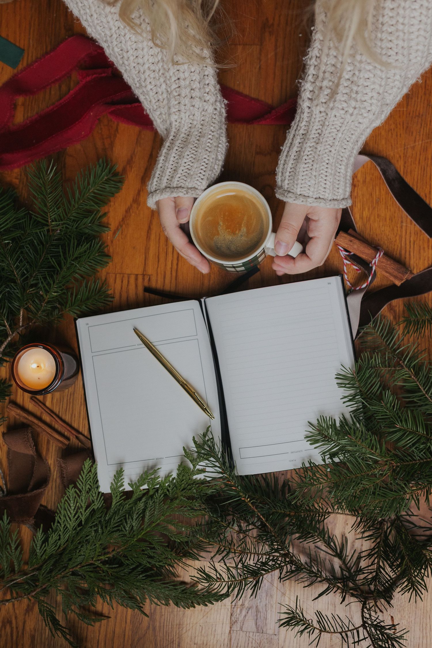 Hands holding coffee cup with notebook and evergreen branches on wooden table.