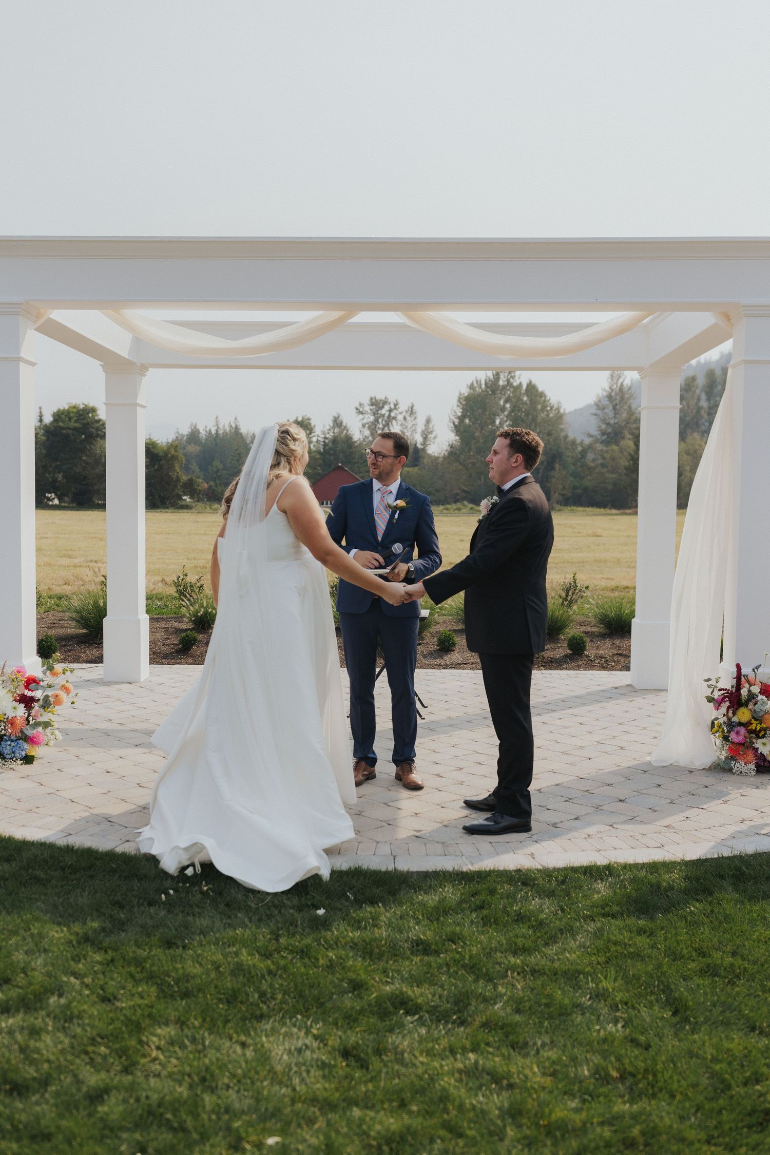 Wedding ceremony under a white columned pergola with floral arrangements.