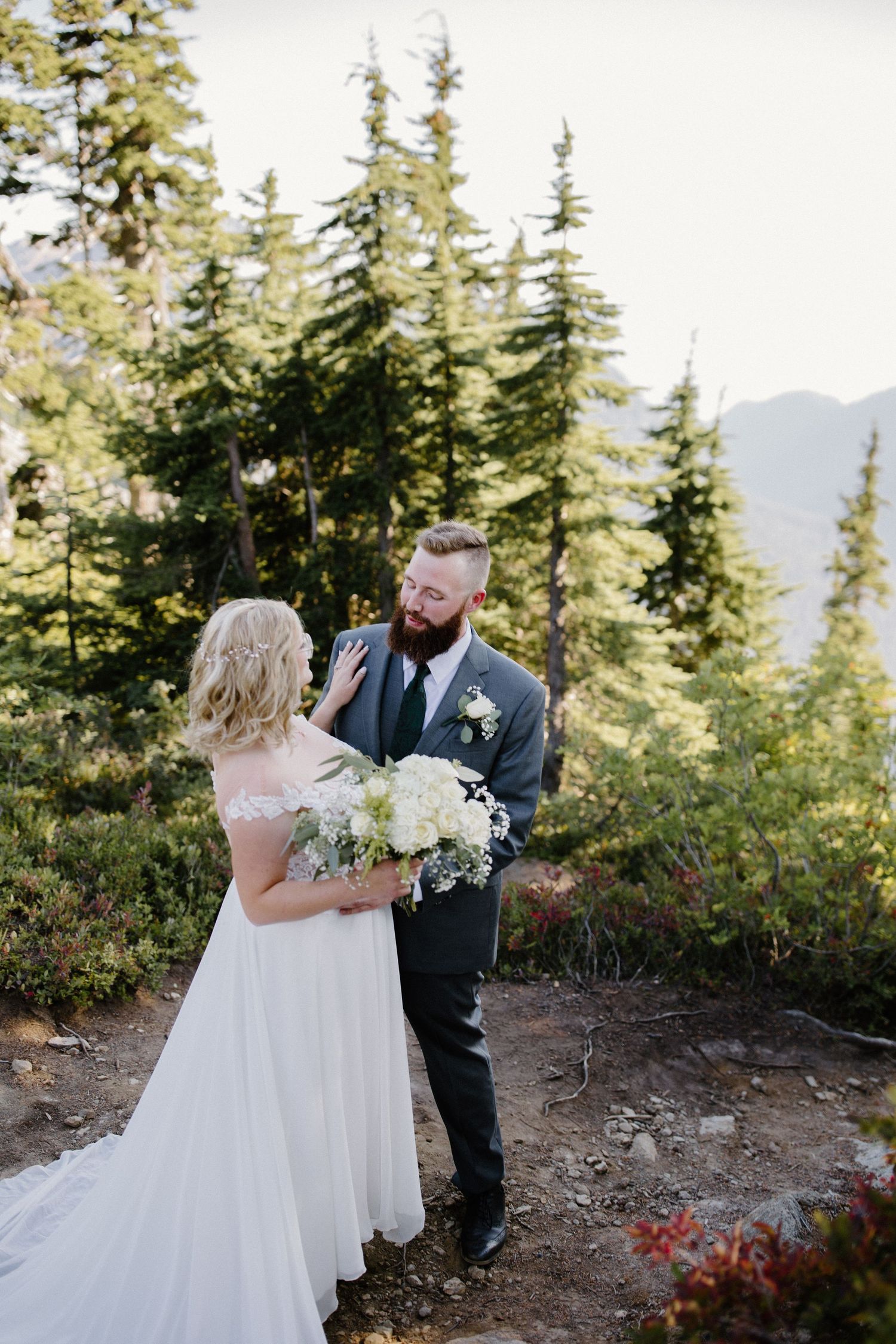 Wedding couple poses among evergreen trees on a mountainside with white floral bouquet.
