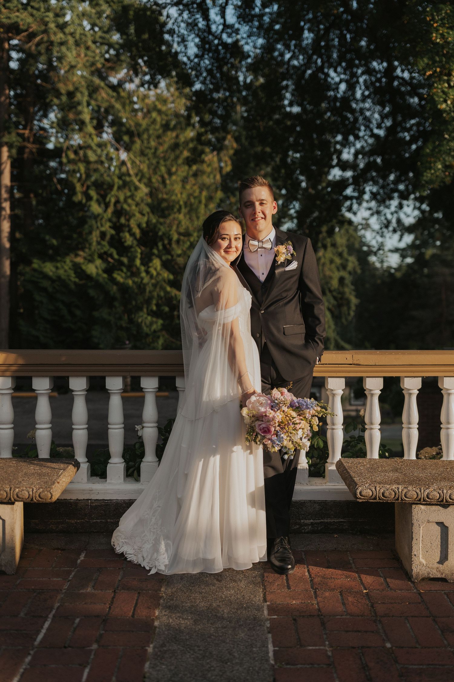 Wedding couple poses on brick terrace with white balustrade during golden hour.