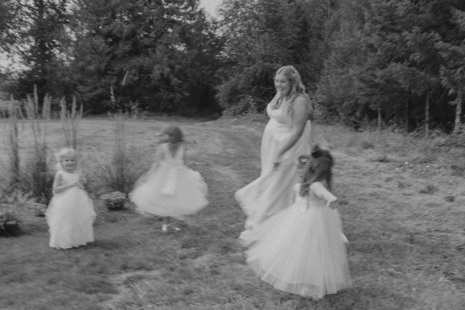 Bridesmaids in flowing white dresses pose together on a grassy hillside with evergreen trees.
