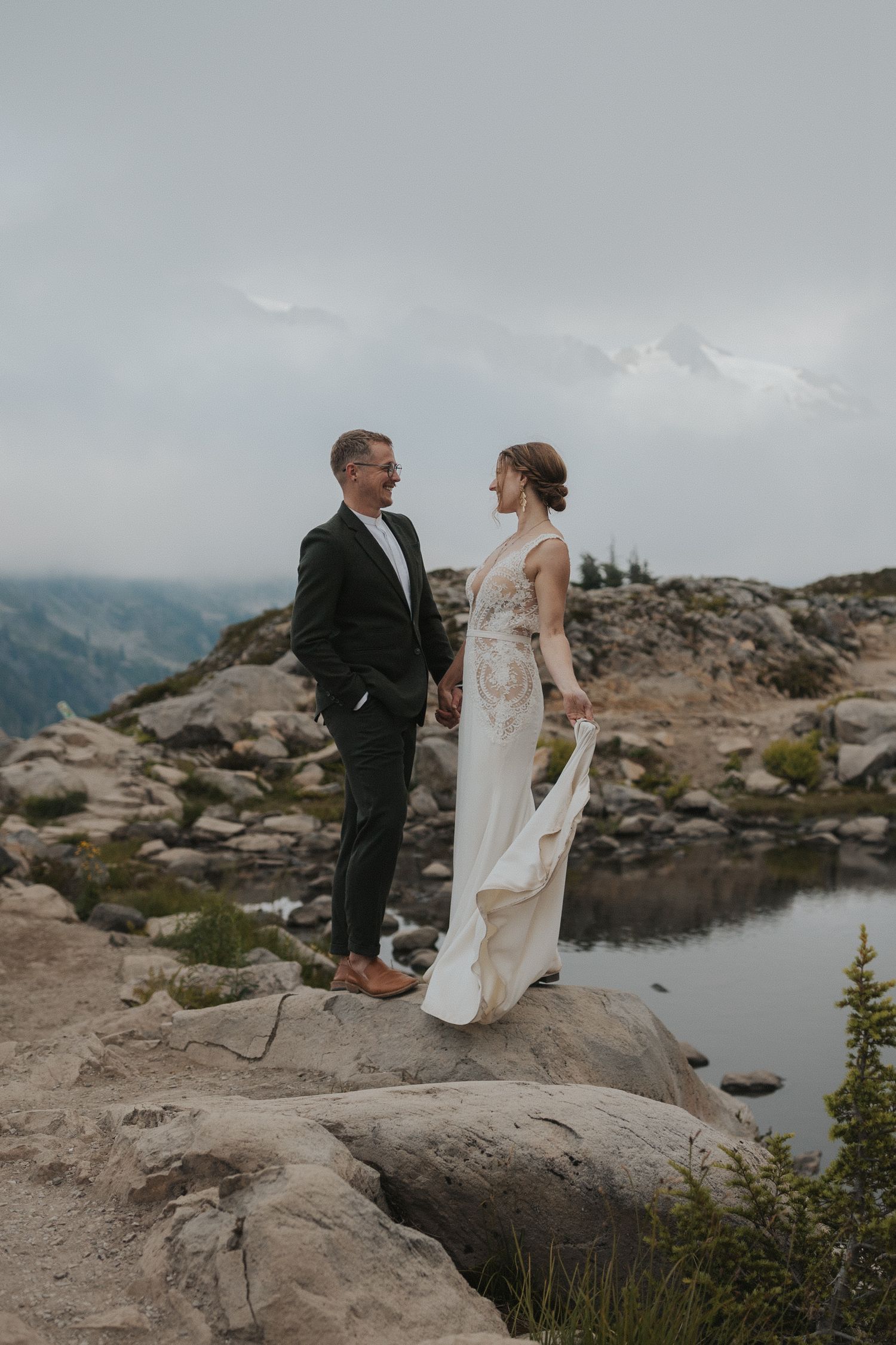 A couple in wedding attire explores a rocky mountain landscape with a small alpine lake in view.