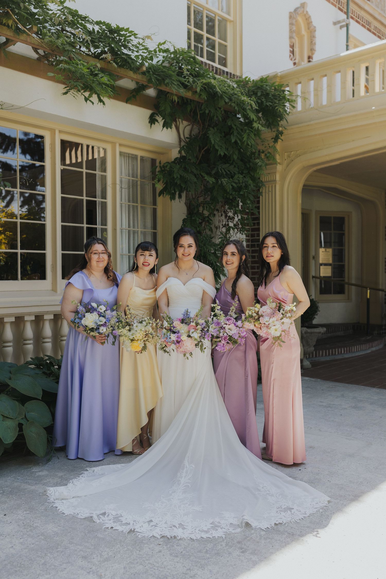 Bridal party in pastel dresses posing with bouquets against classic architectural backdrop.