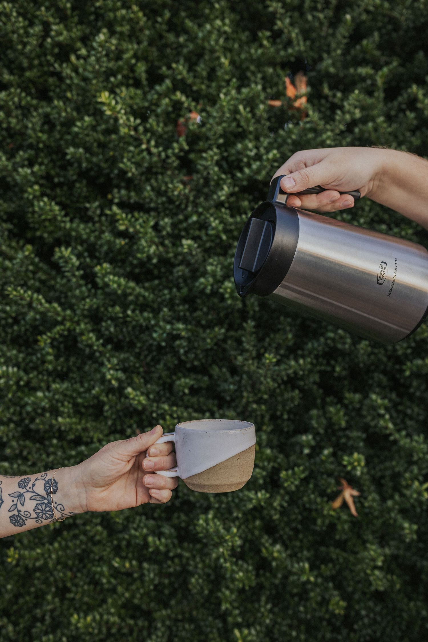 Hands demonstrating the opening of a stainless steel thermos against a green hedge background.