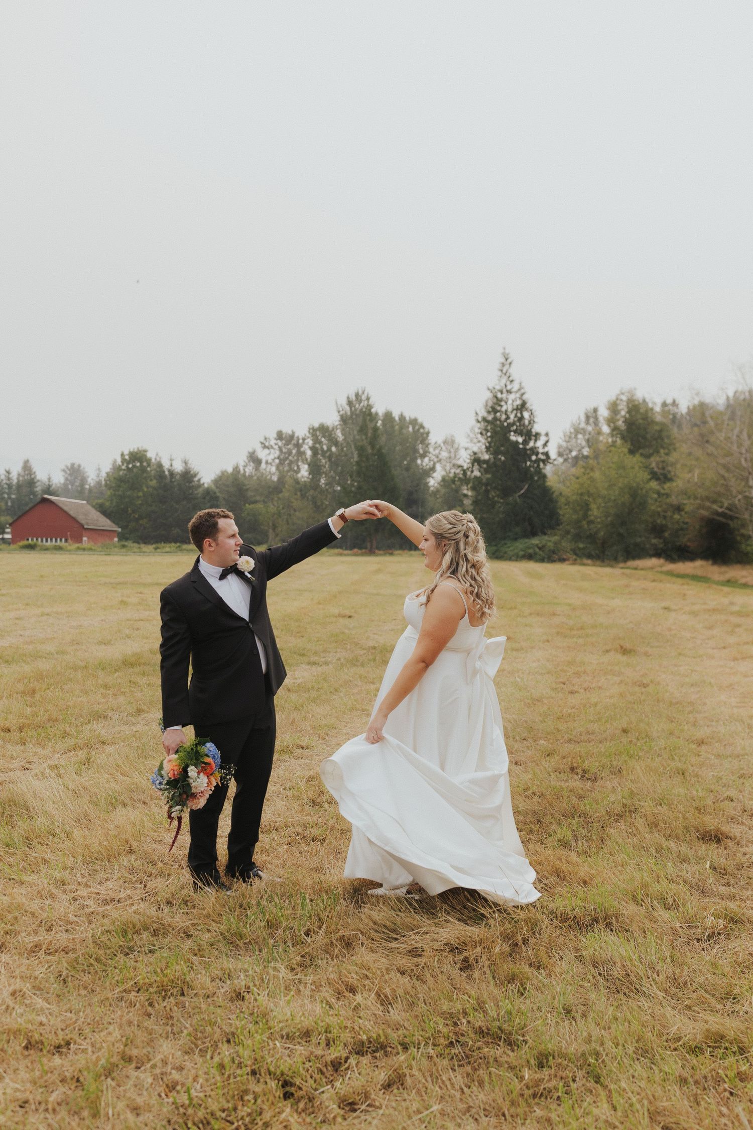 A couple in wedding attire dances playfully on a grassy hillside with a red barn in the distance.