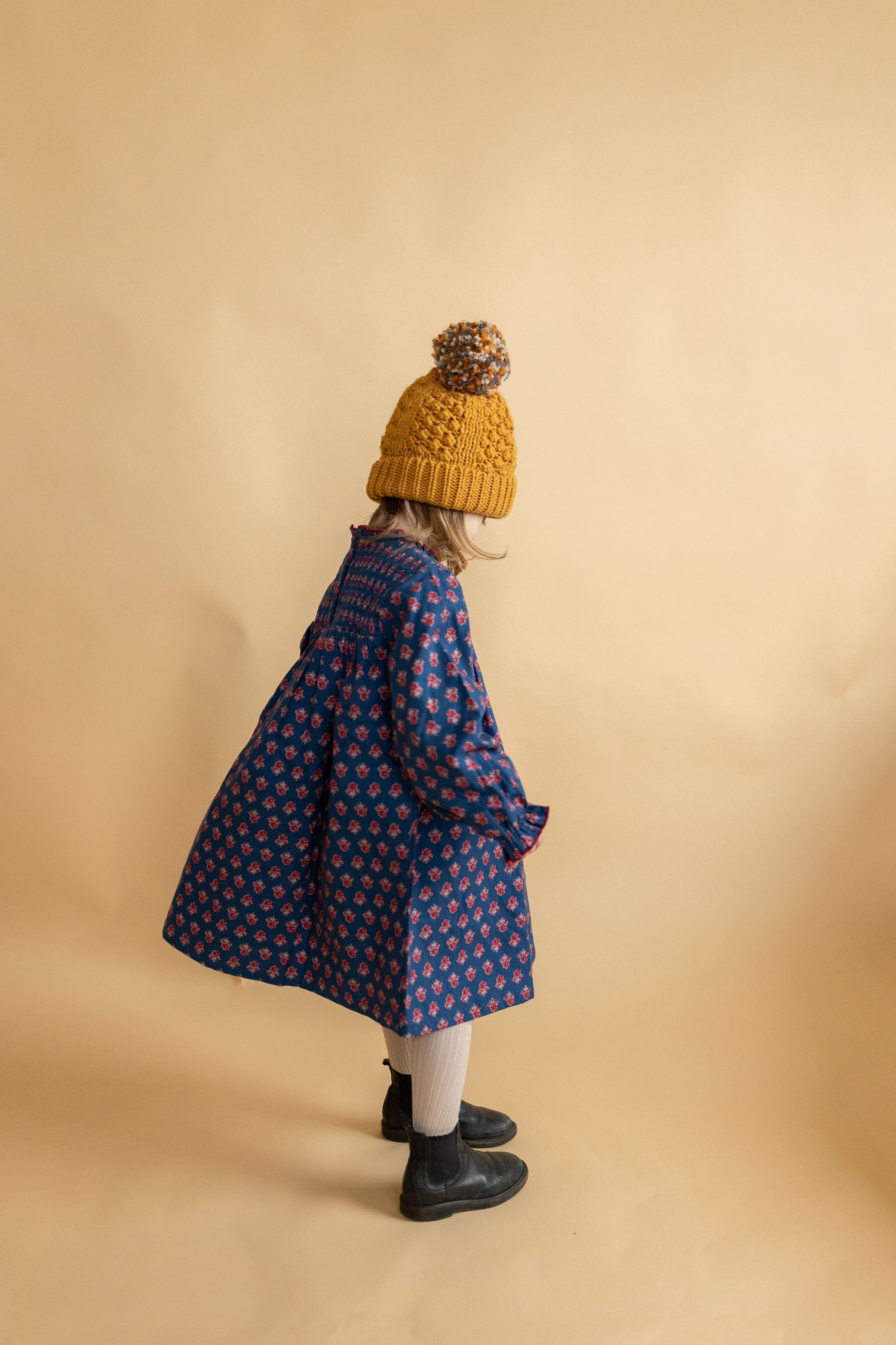 Child wearing blue polka dot dress and yellow beanie against beige background.