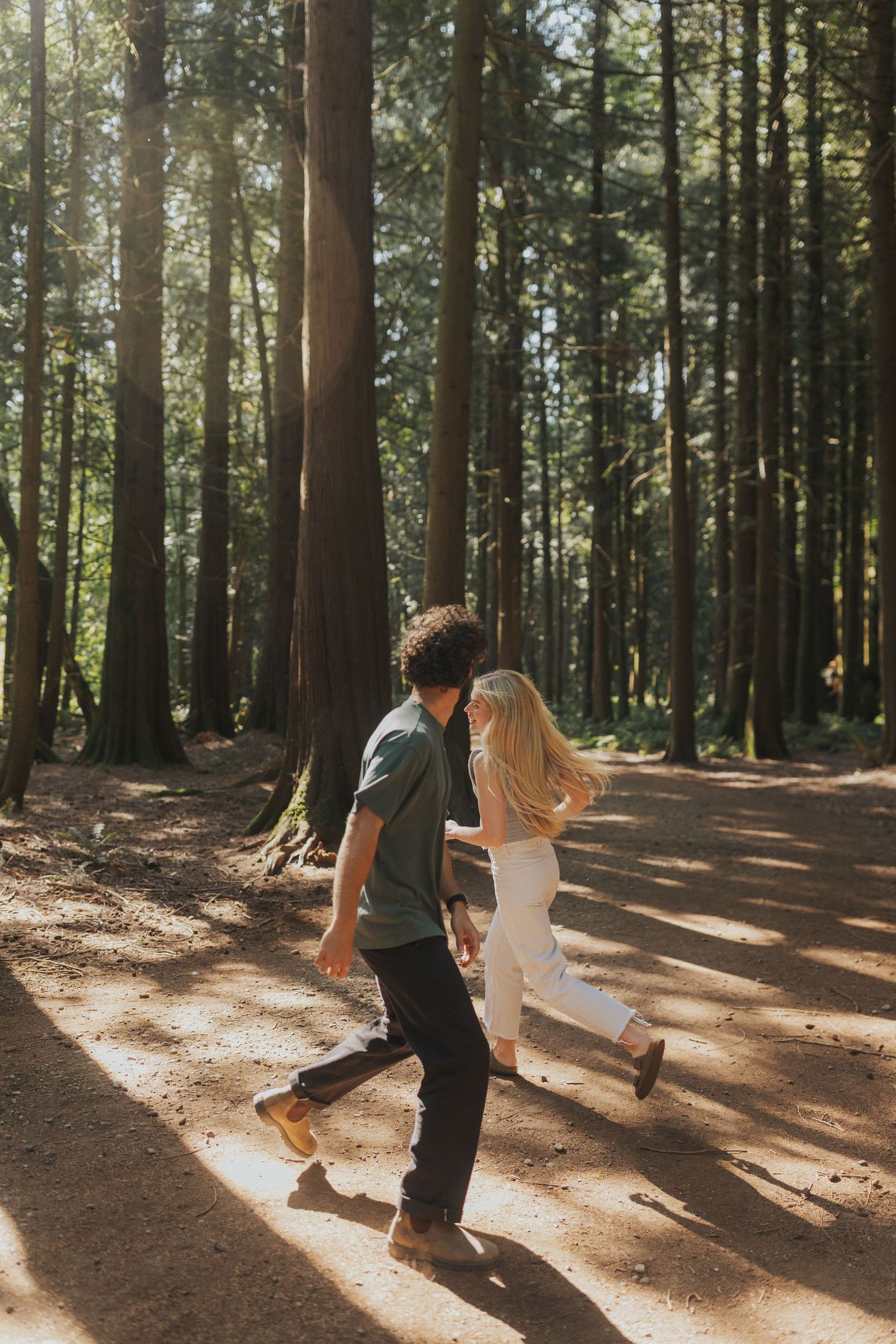 A couple dances playfully on a forest path with sunbeams streaming through the trees.