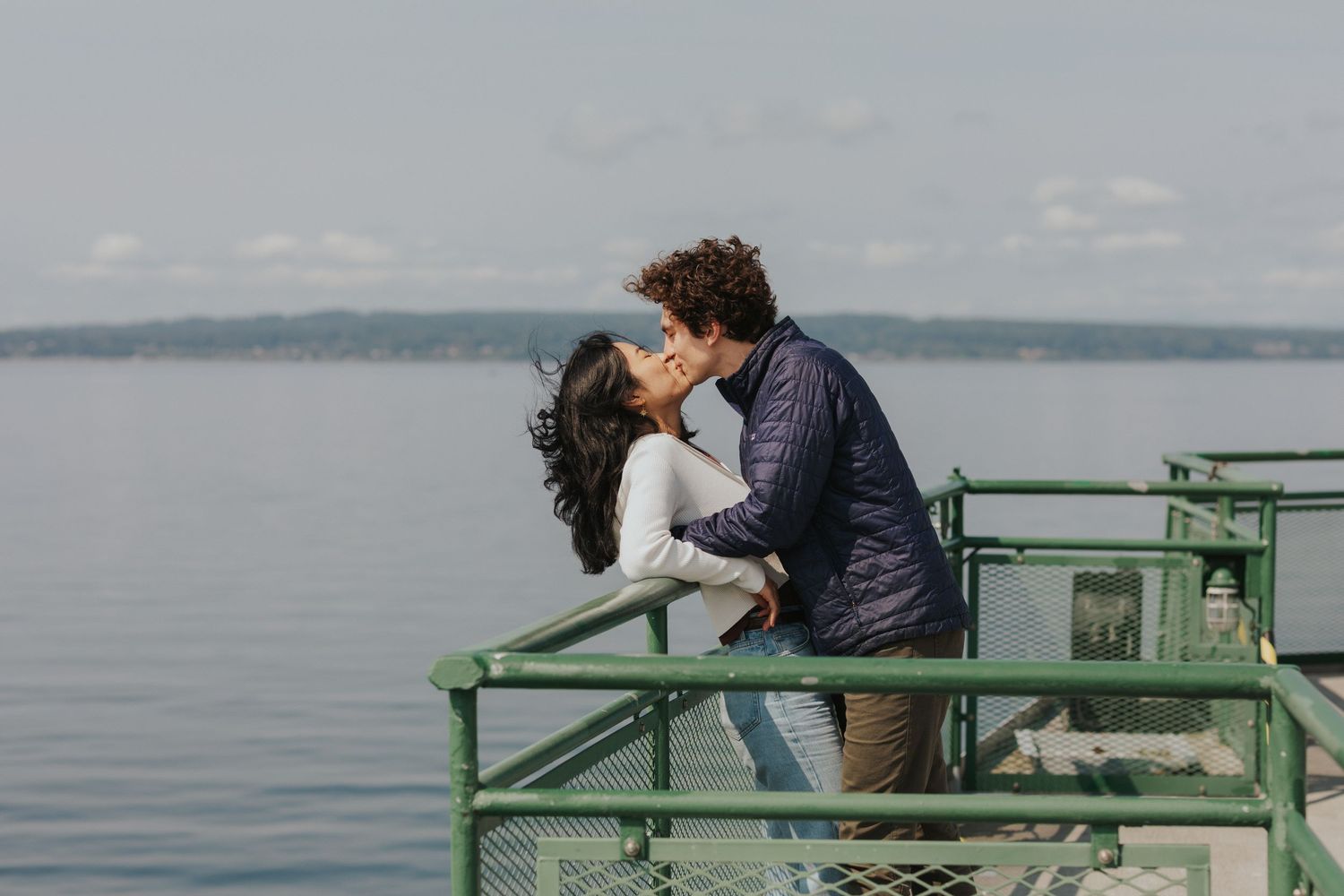 A couple embraces on a green-railed deck against a tranquil ocean backdrop.