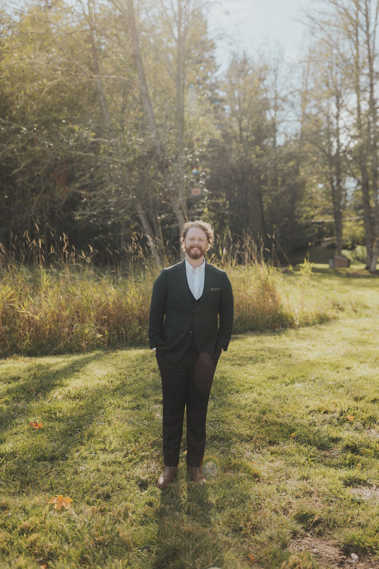 Groom in classic black suit stands alone in a sunlit autumn field.