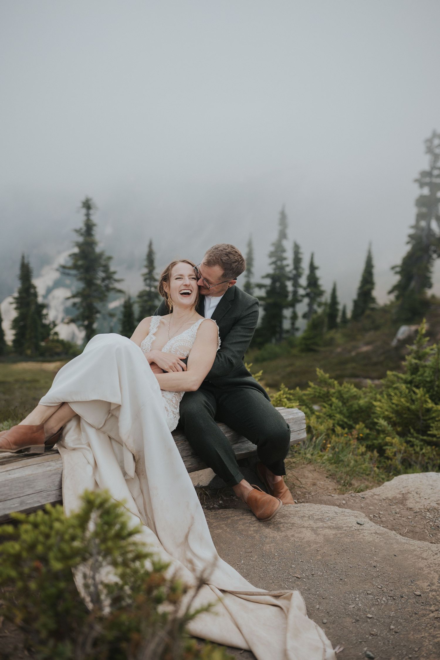 Playful wedding couple laughs while sitting on rocky mountain ledge surrounded by pine trees.