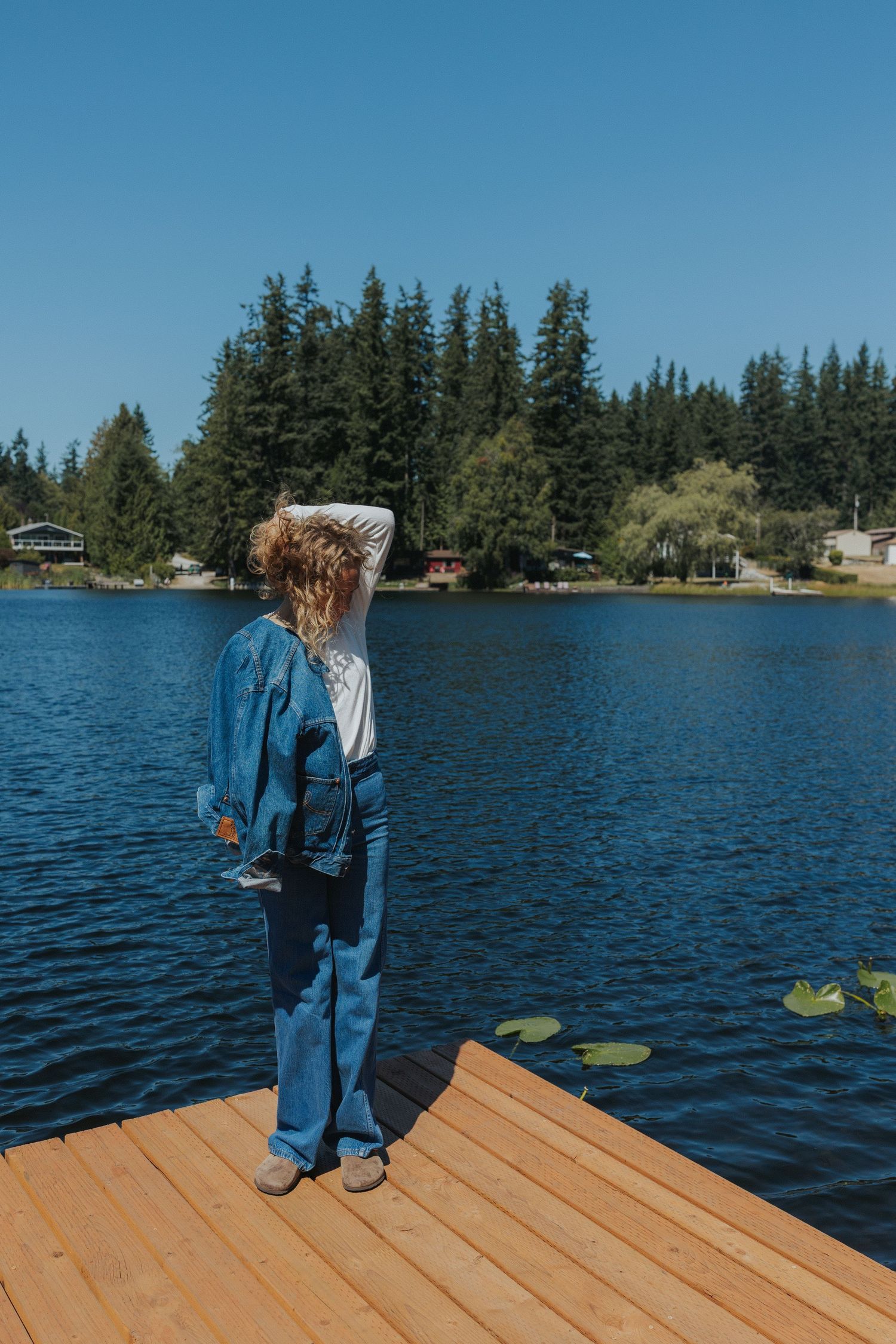 A person in denim poses energetically on a lakeside dock with evergreen trees in the distance.