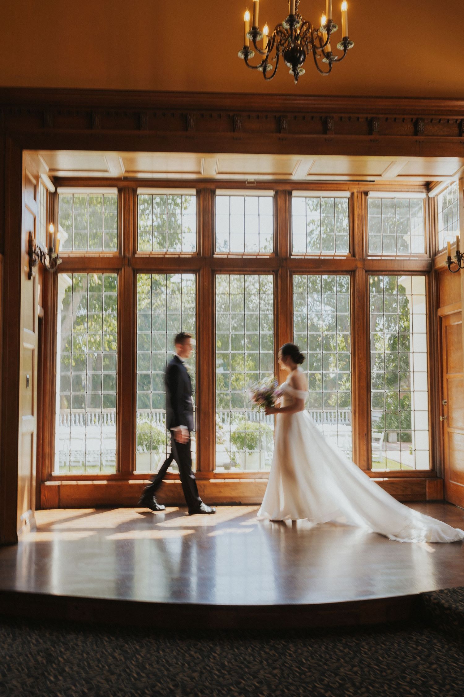 Elegant wedding scene in a grand room with large windows and chandelier overhead.