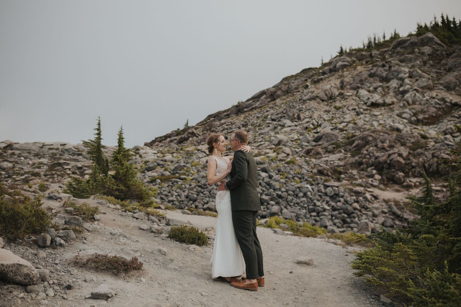Two people walk along a rugged mountain trail surrounded by rocky terrain and evergreen trees.