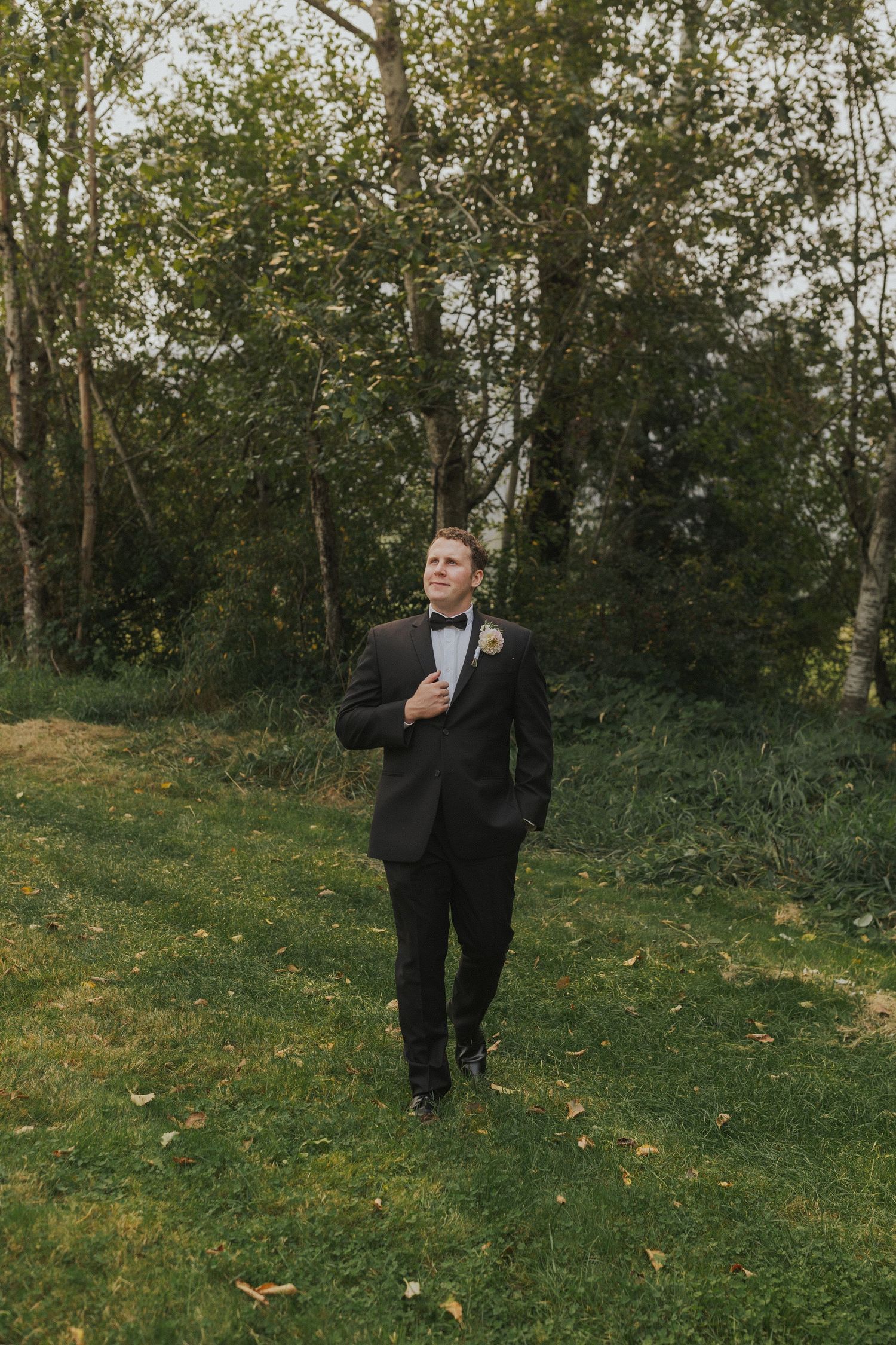 A groom in a black tuxedo stands thoughtfully on a grassy slope with trees in the background.