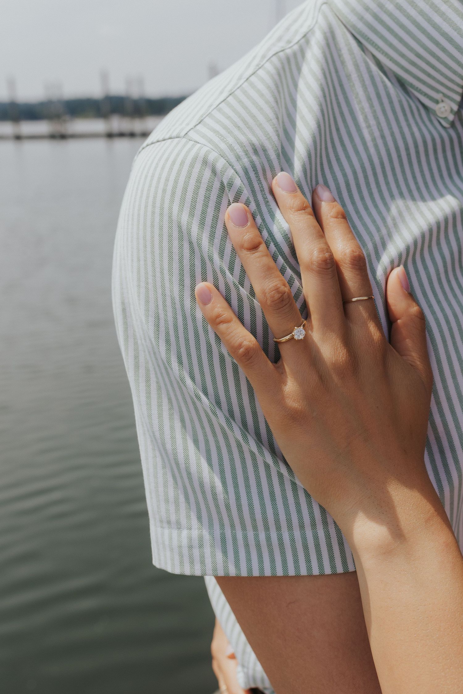 Close-up of hands showing off an engagement ring against a striped fabric with water in the background.