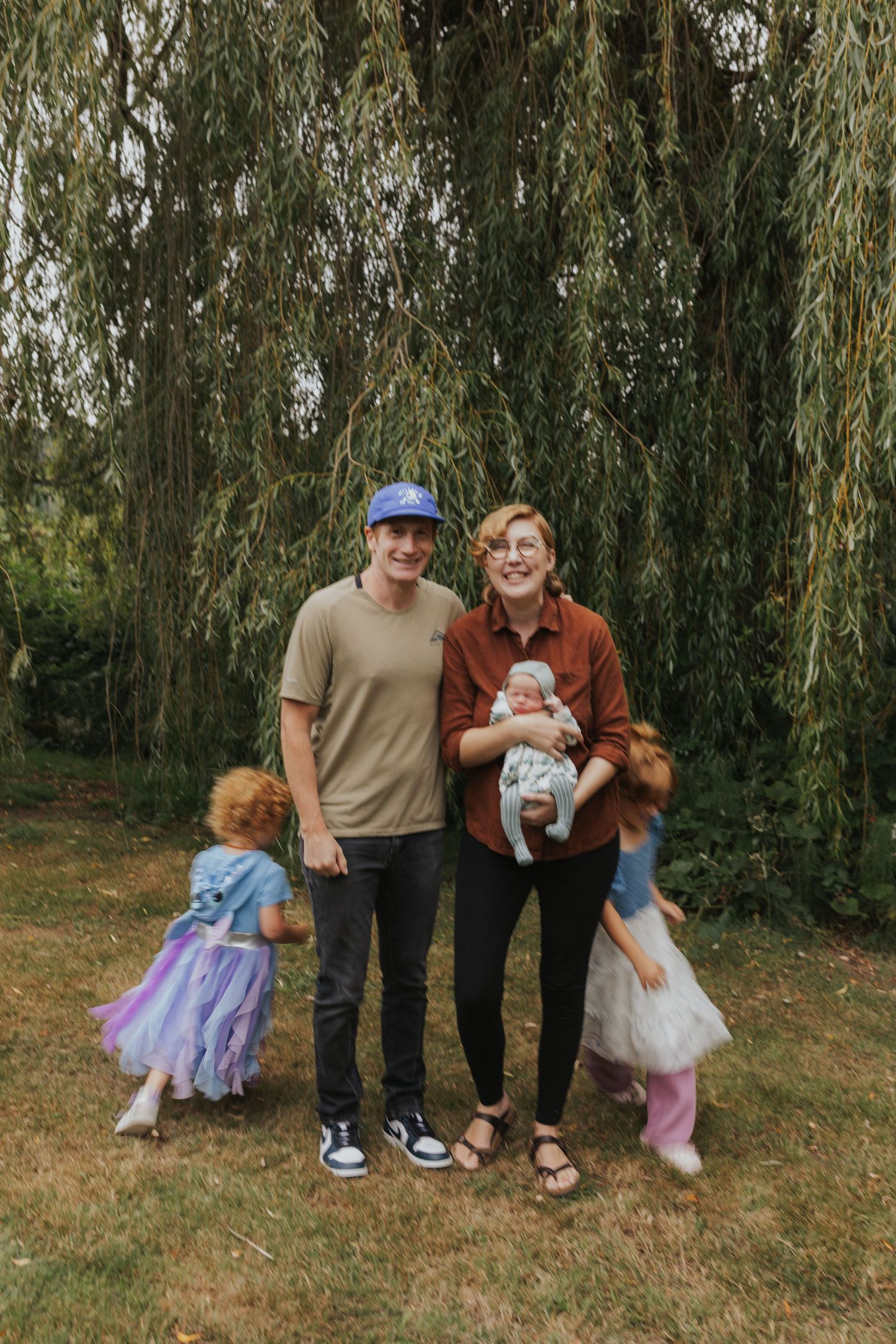 Family portrait under weeping willow trees with children in whimsical outfits.