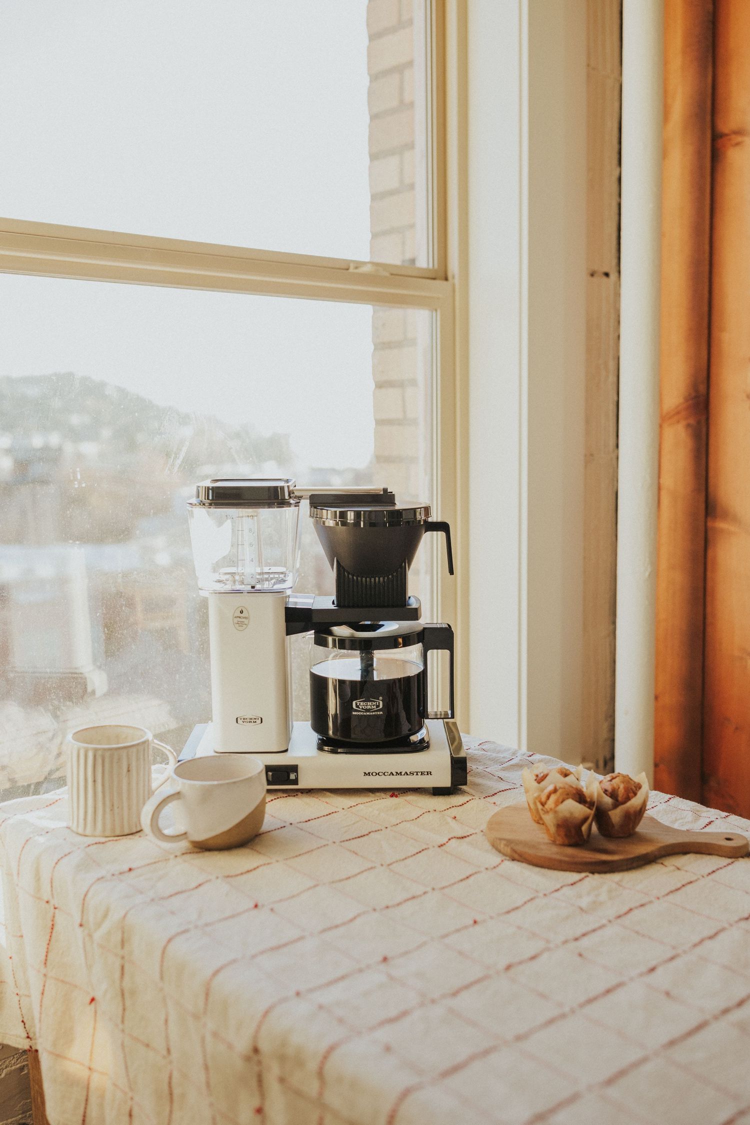Coffee maker and pastries on tablecloth by window with mountain view.
