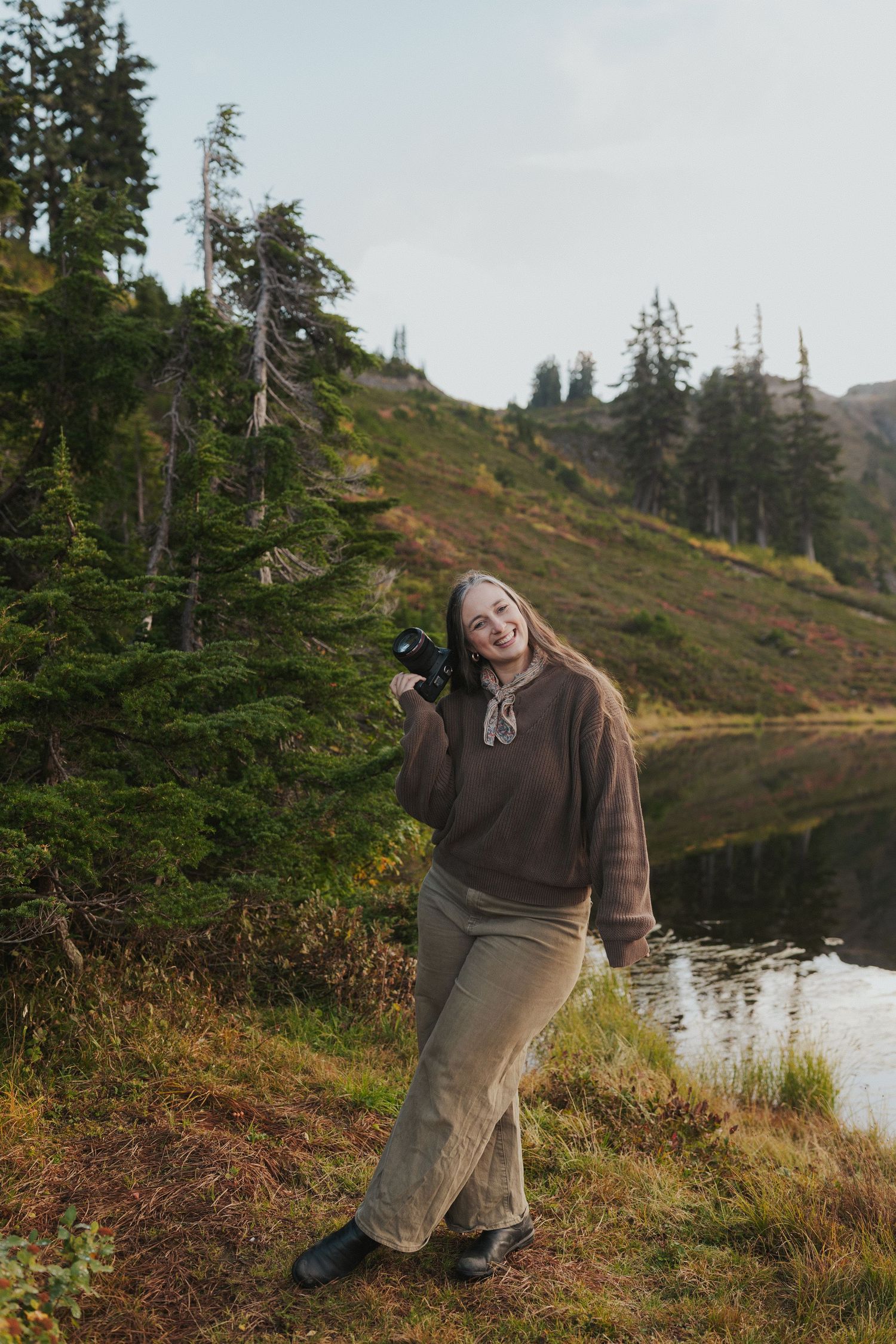 Nature photographer in casual attire poses with camera near mountain lake and evergreen trees.