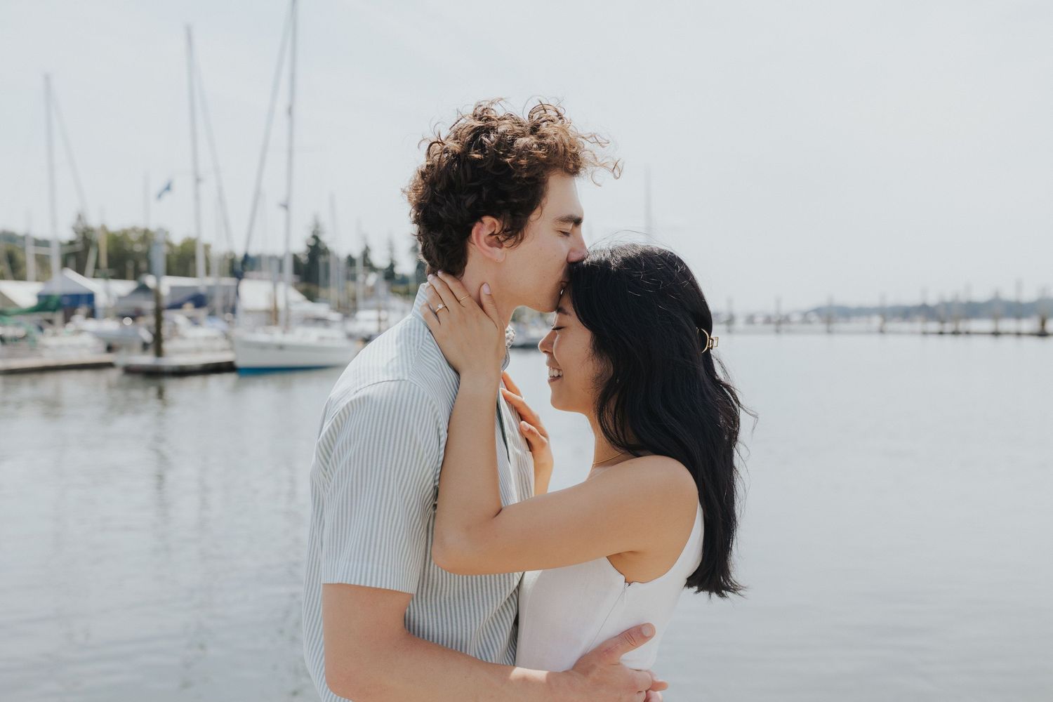 Couple embracing by the waterfront with boats and docks visible in the distance.