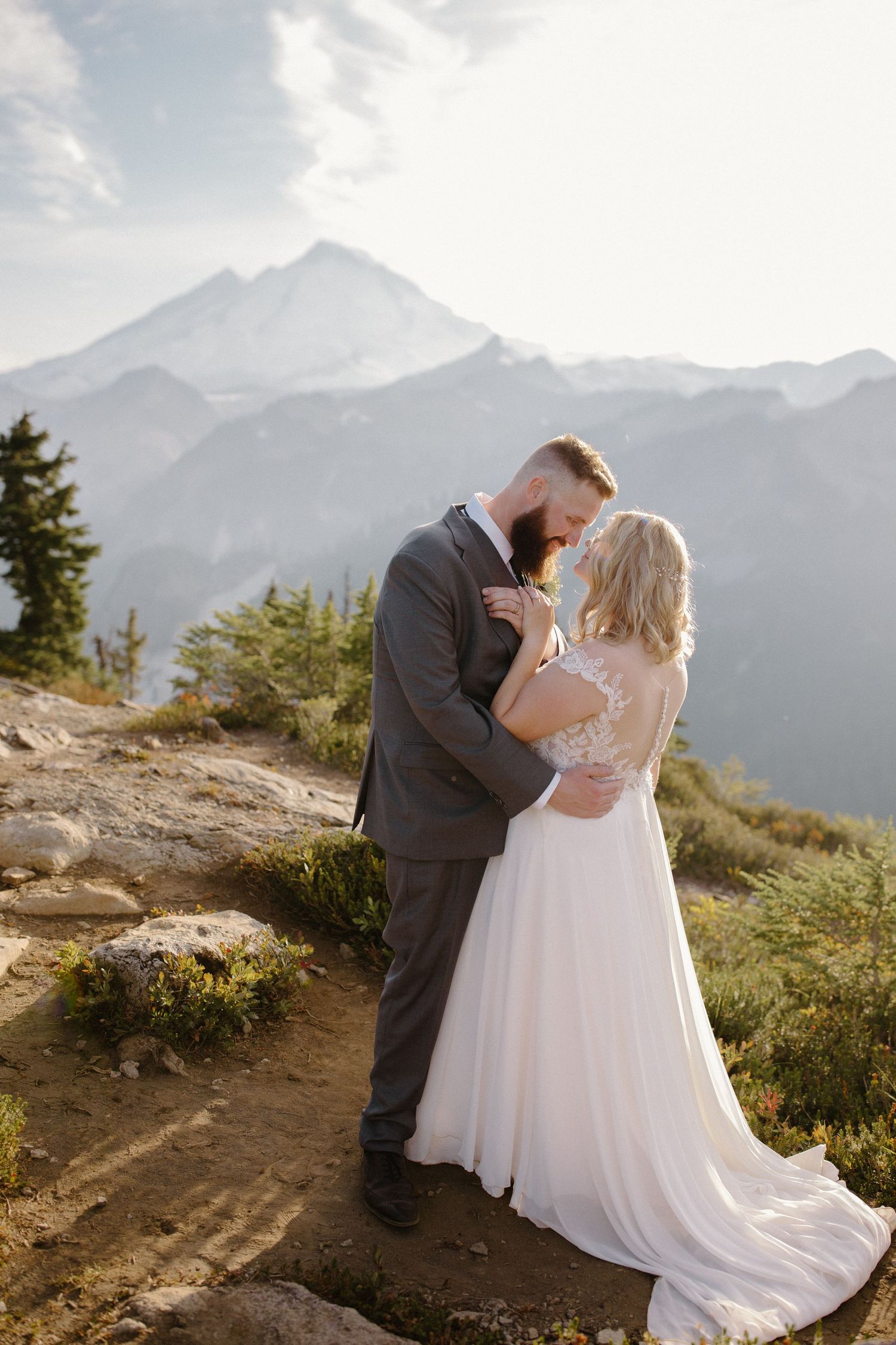 Newlyweds embrace on a mountain trail with majestic peaks and evergreen trees in the background.