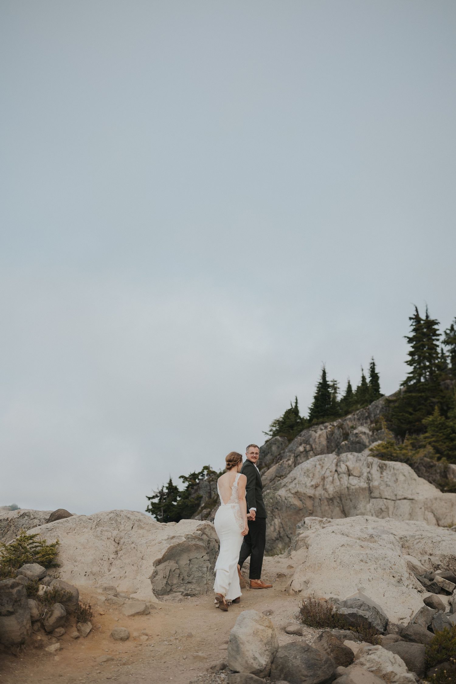 Two people in wedding attire navigate across a rocky mountain outcropping against an overcast sky.