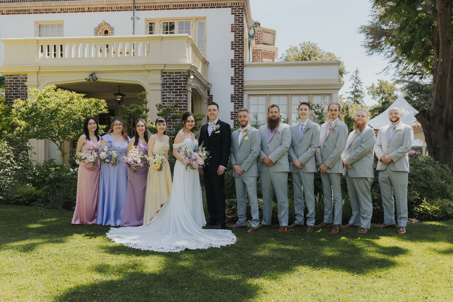 Wedding party in pastel attire posed together on lawn in front of historic mansion.
