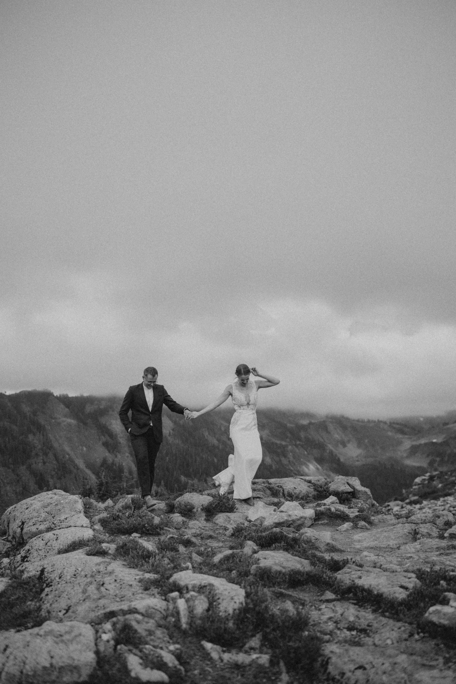 Two people in wedding attire dance on a dramatic mountain summit with cloudy skies.