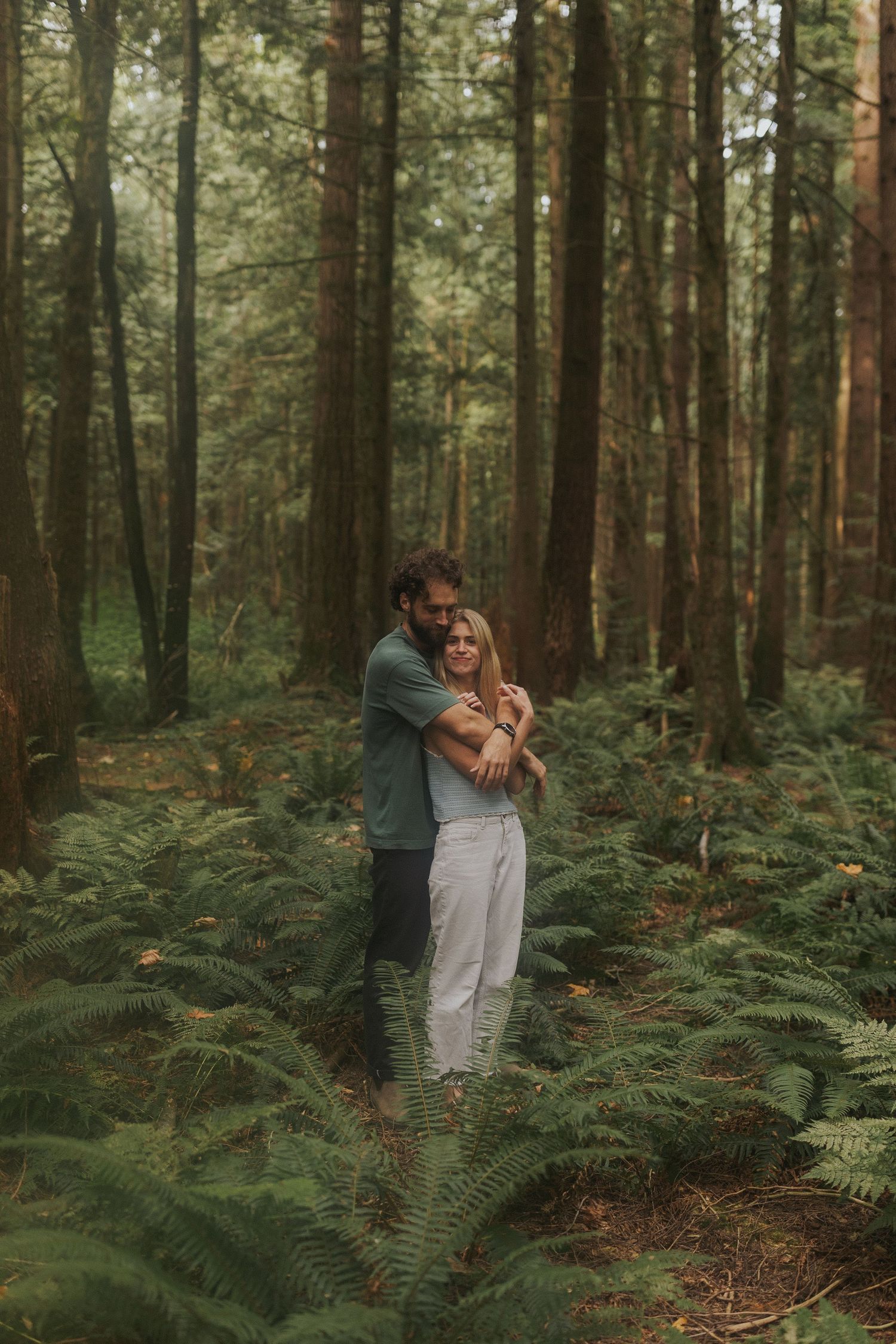 A romantic couple embraces while standing among tall evergreen trees and ferns in a misty forest.