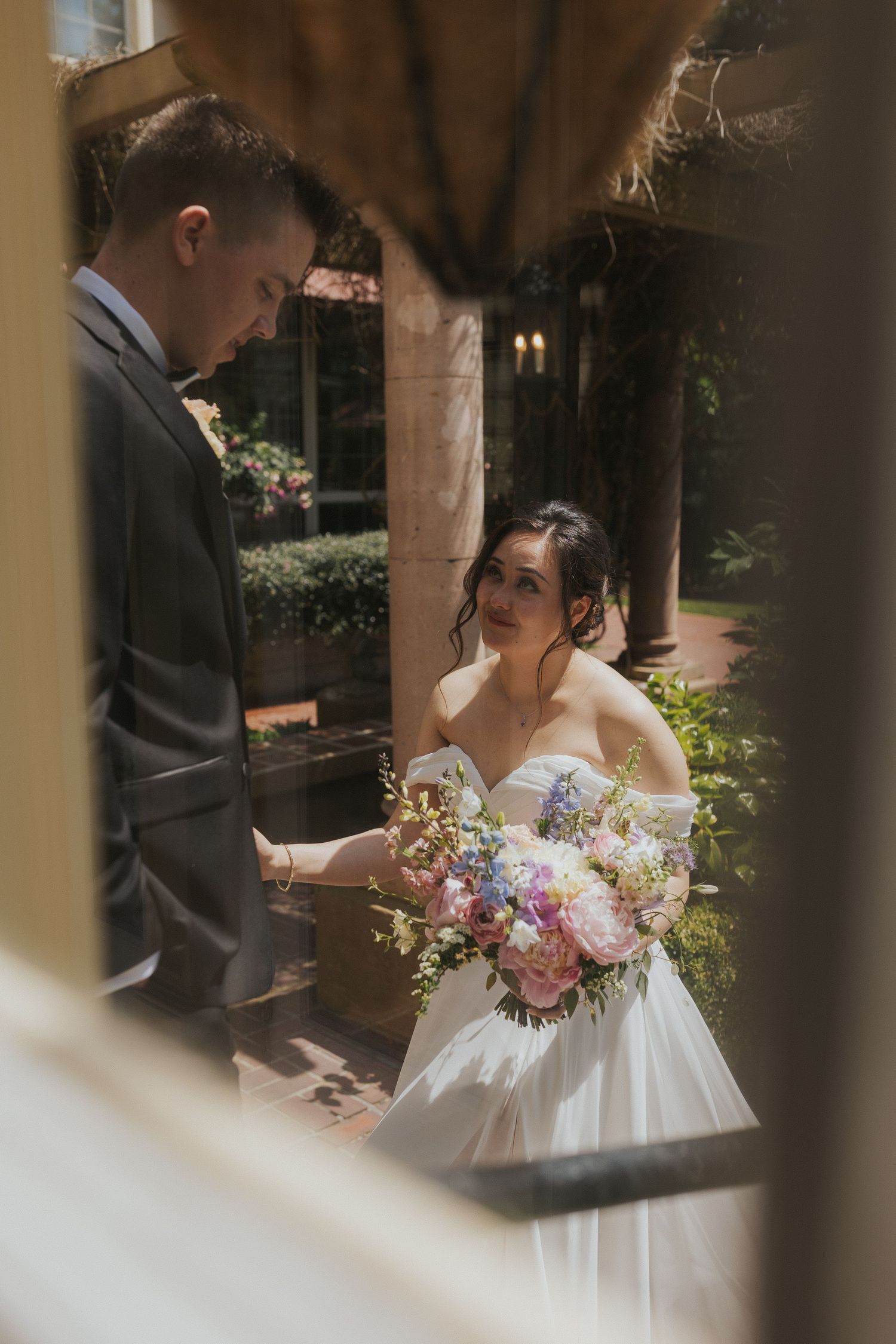 Intimate wedding moment captured through a window frame with a couple exchanging vows and a beautiful pastel bouquet.