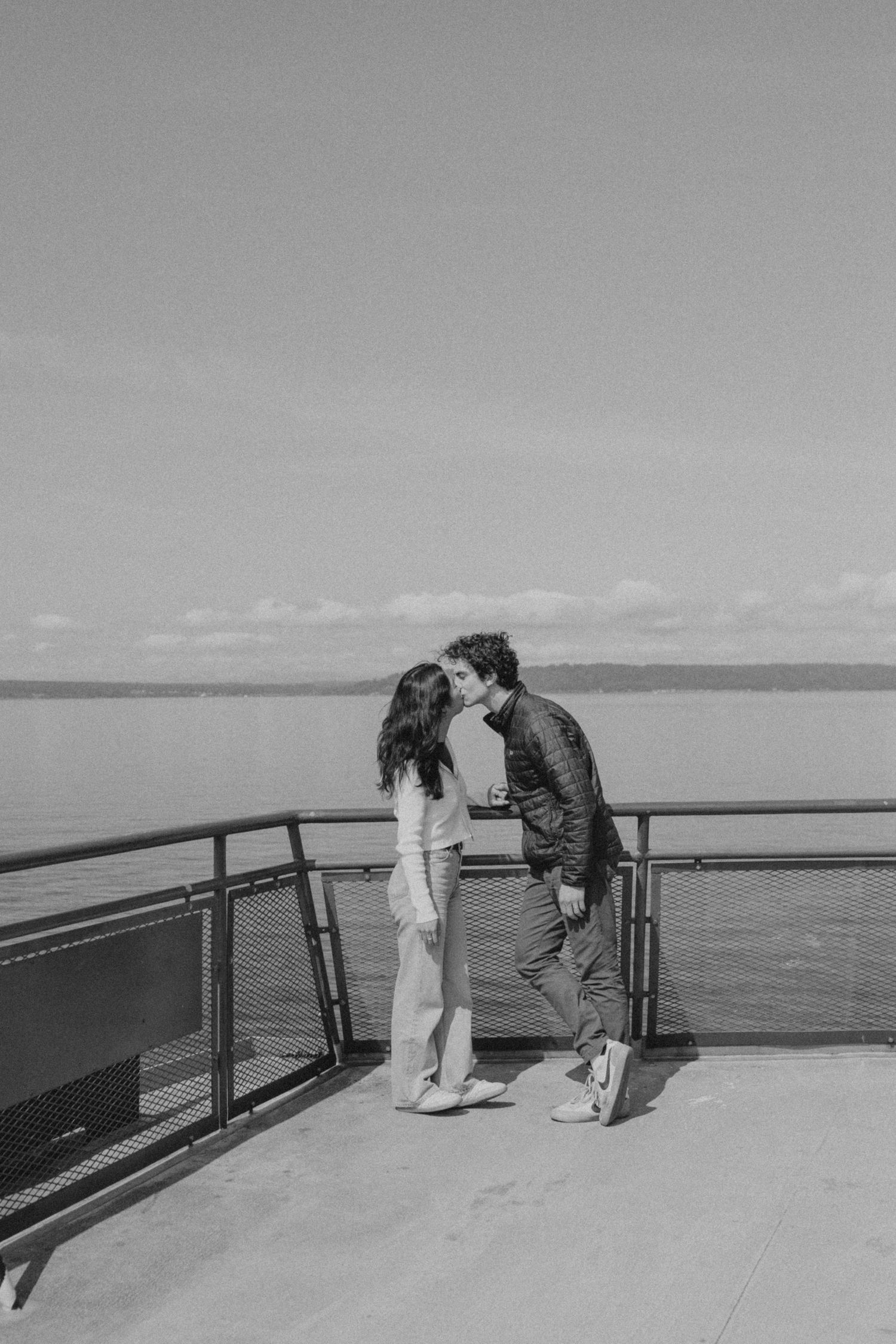 A couple shares a tender moment on a viewing platform overlooking a serene waterfront on a cloudy day.