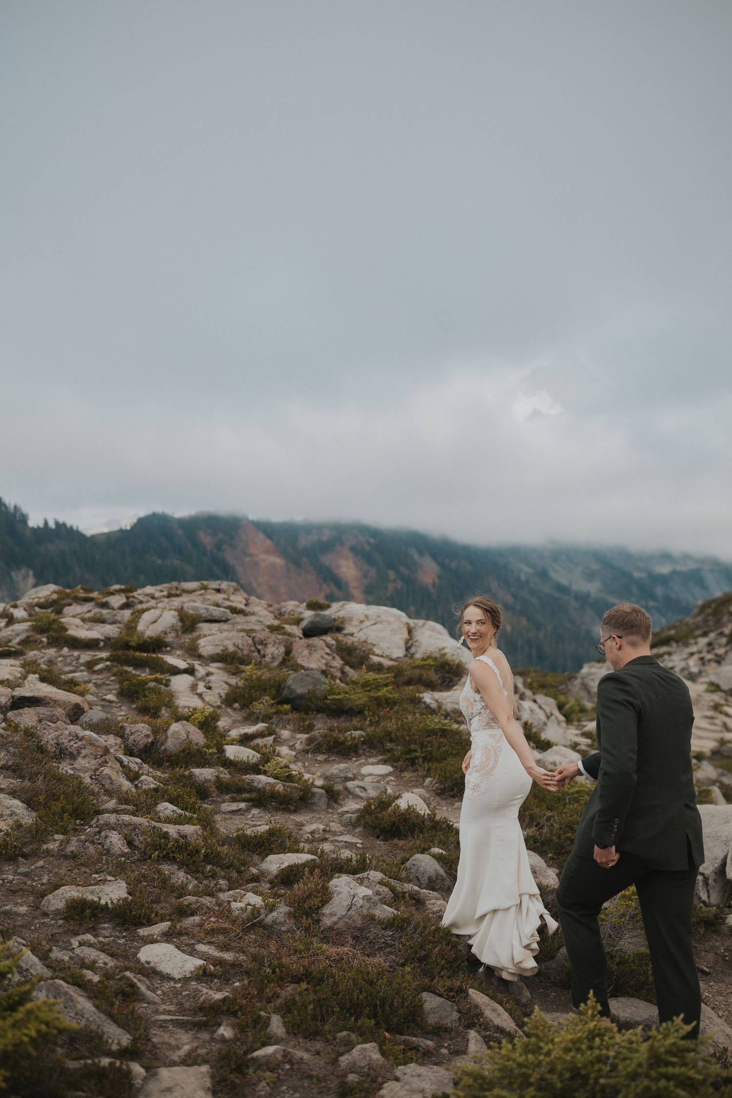 A couple traverses rocky mountain terrain with fog-covered peaks in the background.