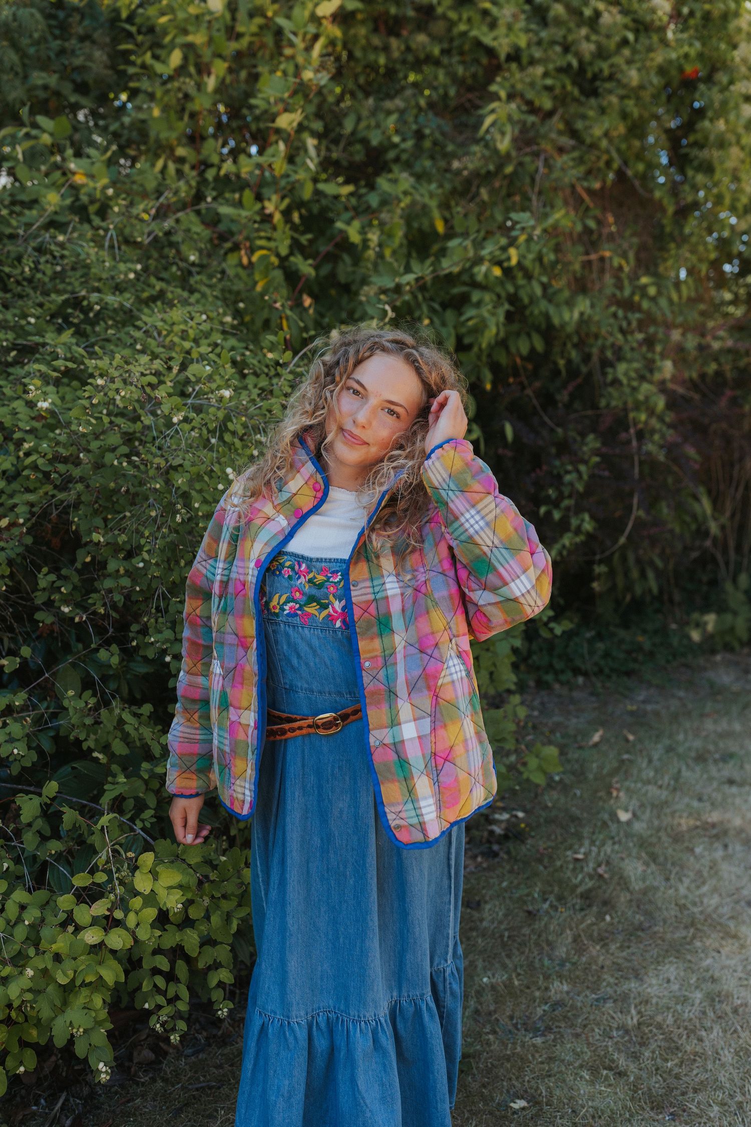 A fashion shoot outdoors featuring a denim dress paired with a colorful plaid jacket against green foliage.