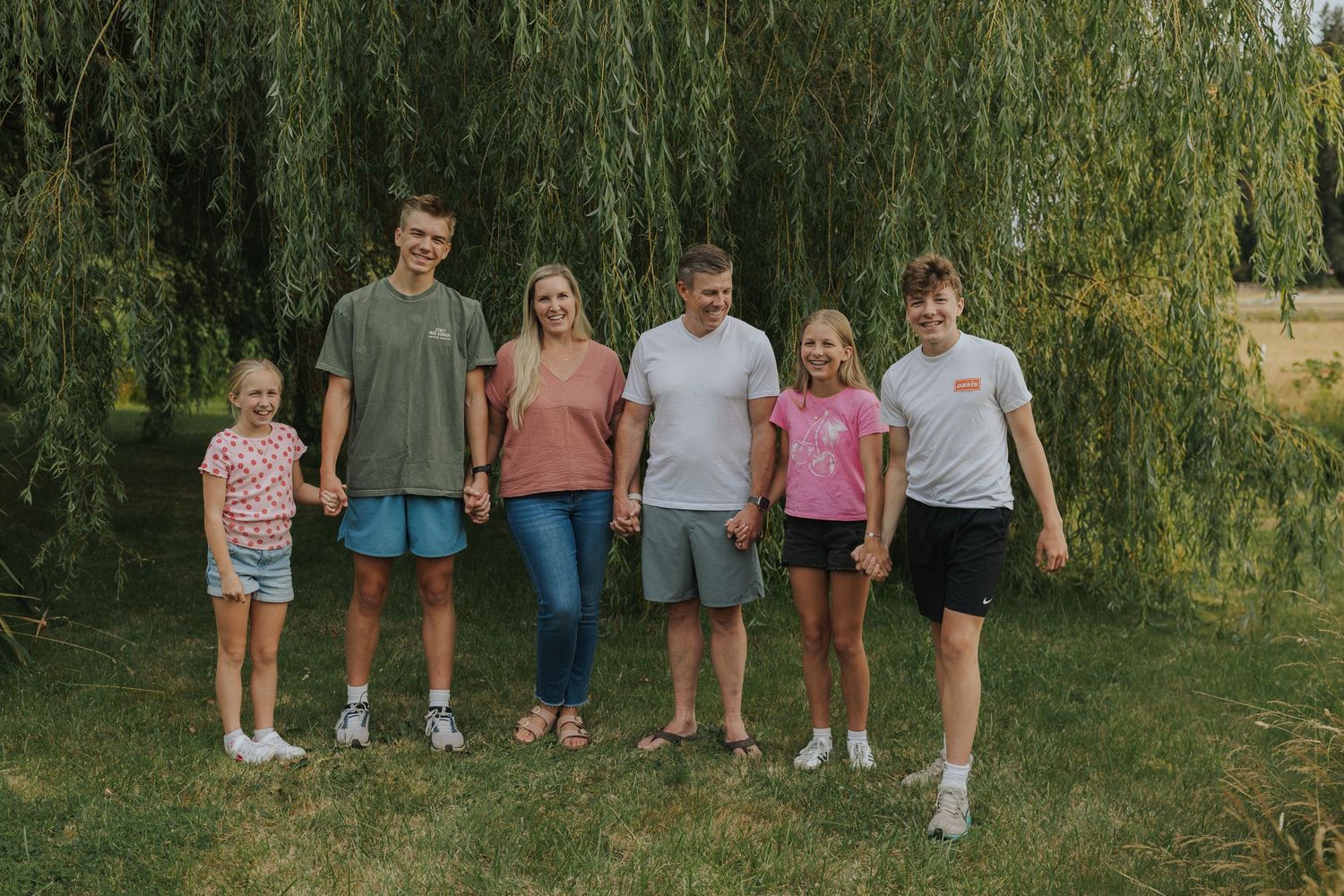 Extended family lined up for casual outdoor portrait beneath willow trees.