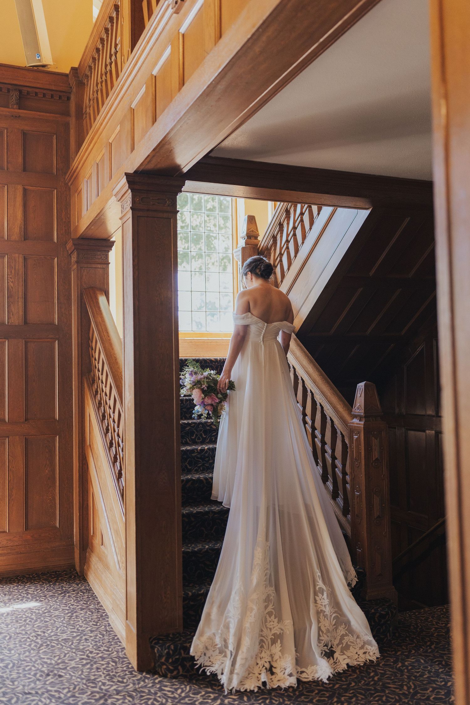 Bride in flowing lace gown ascending wooden staircase in historic venue.