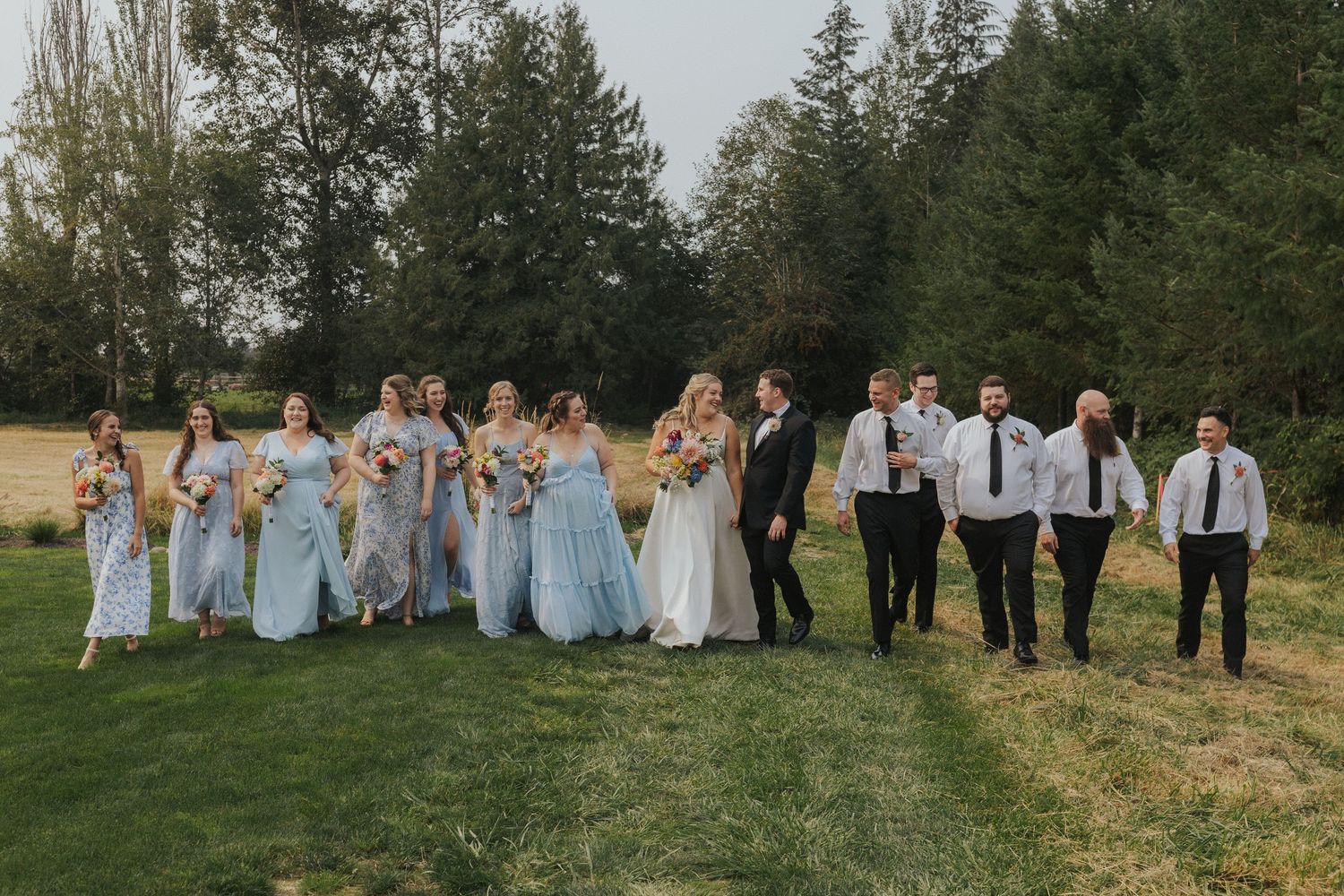 A wedding party in light blue dresses and dark suits poses together on a grassy hillside.