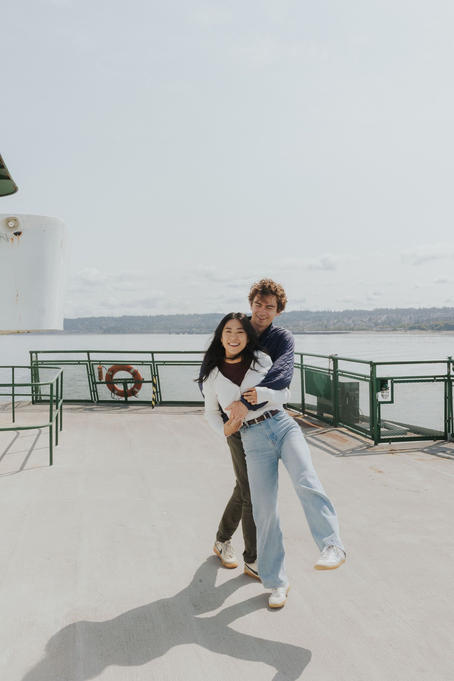 A playful couple embraces on the deck of a ferry with water and mountains in the background.