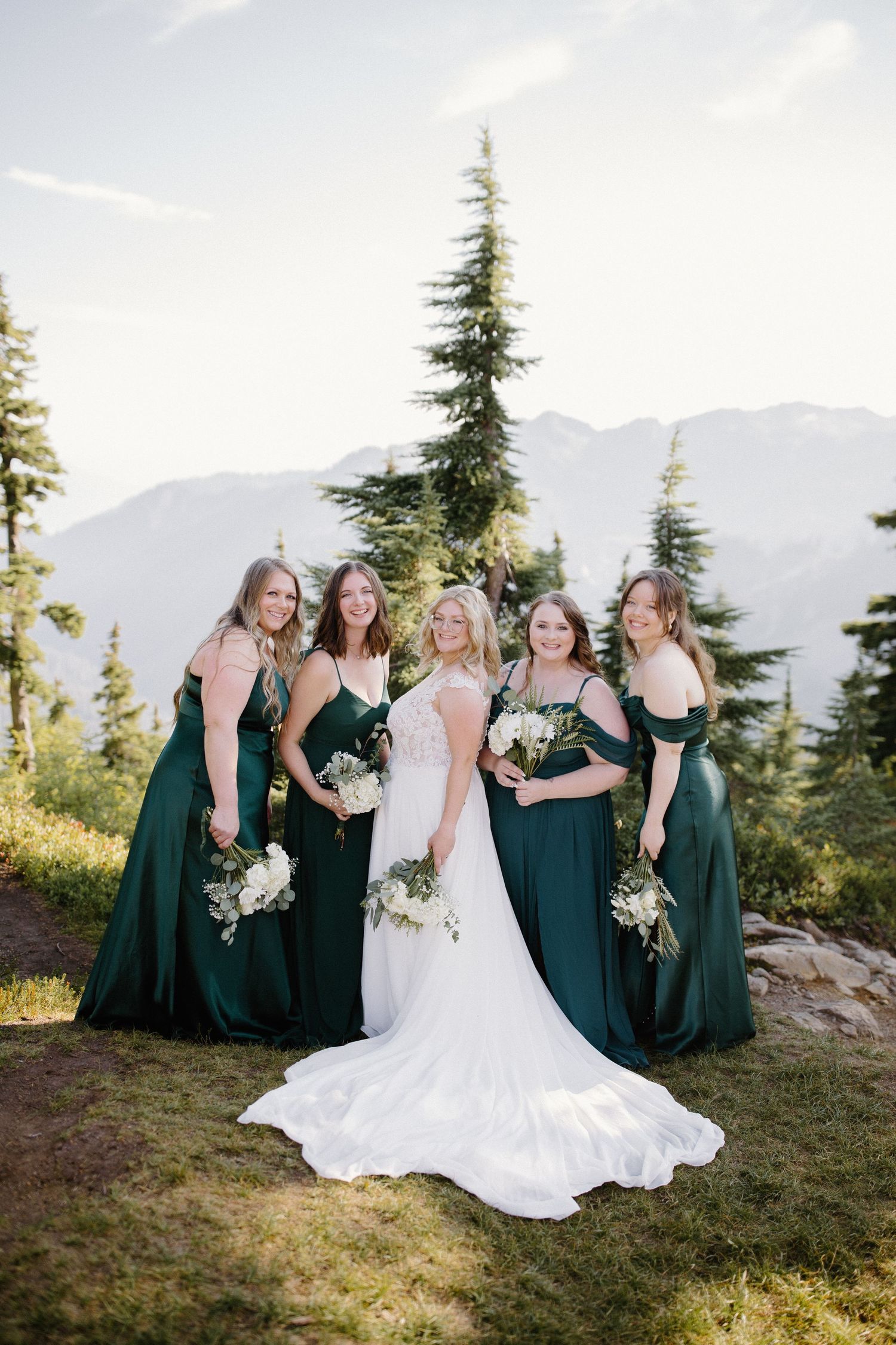 Wedding party in emerald green dresses poses together on a scenic mountain overlook.