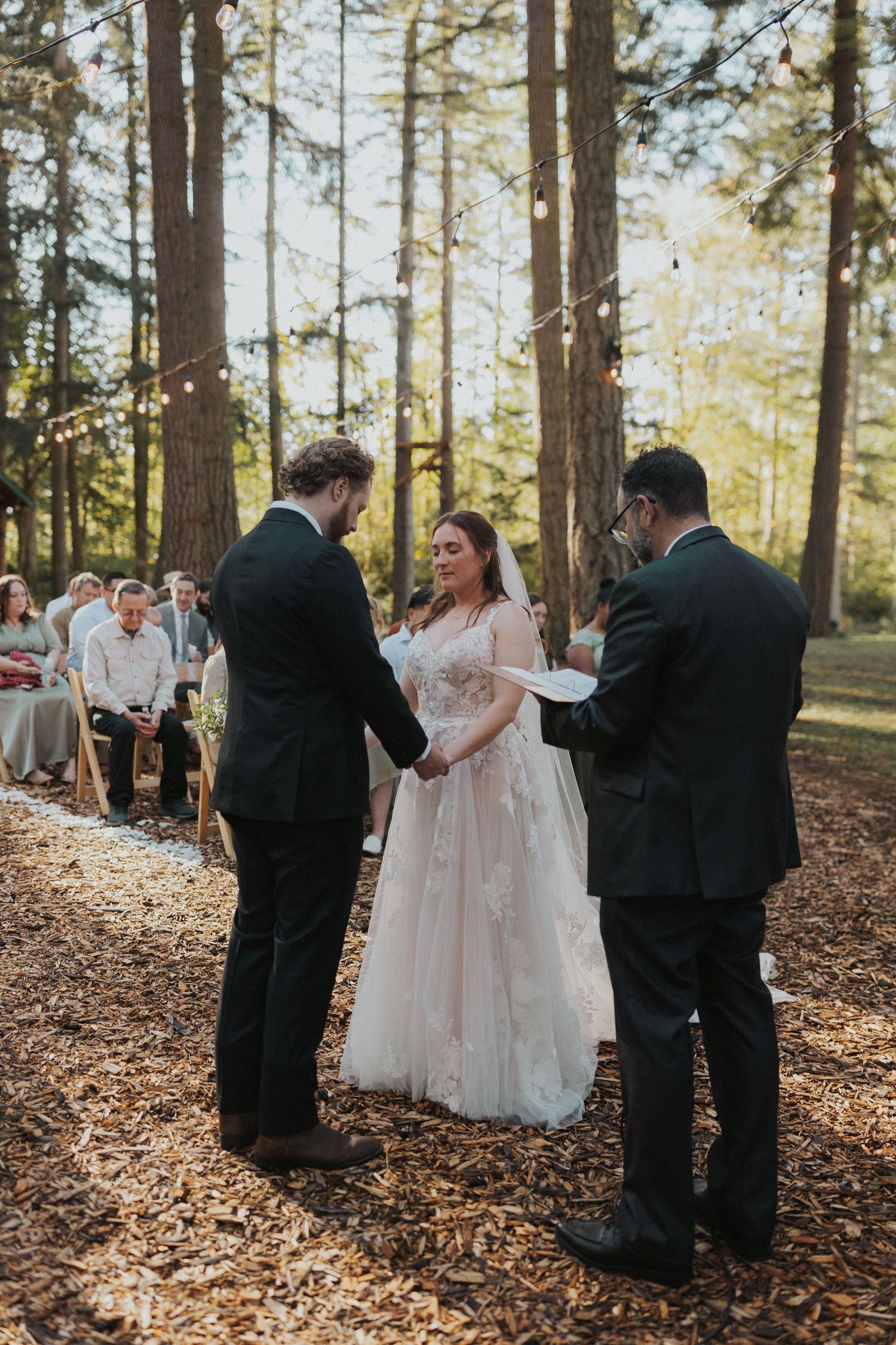 A wedding ceremony taking place on a leaf-covered forest floor surrounded by towering trees.