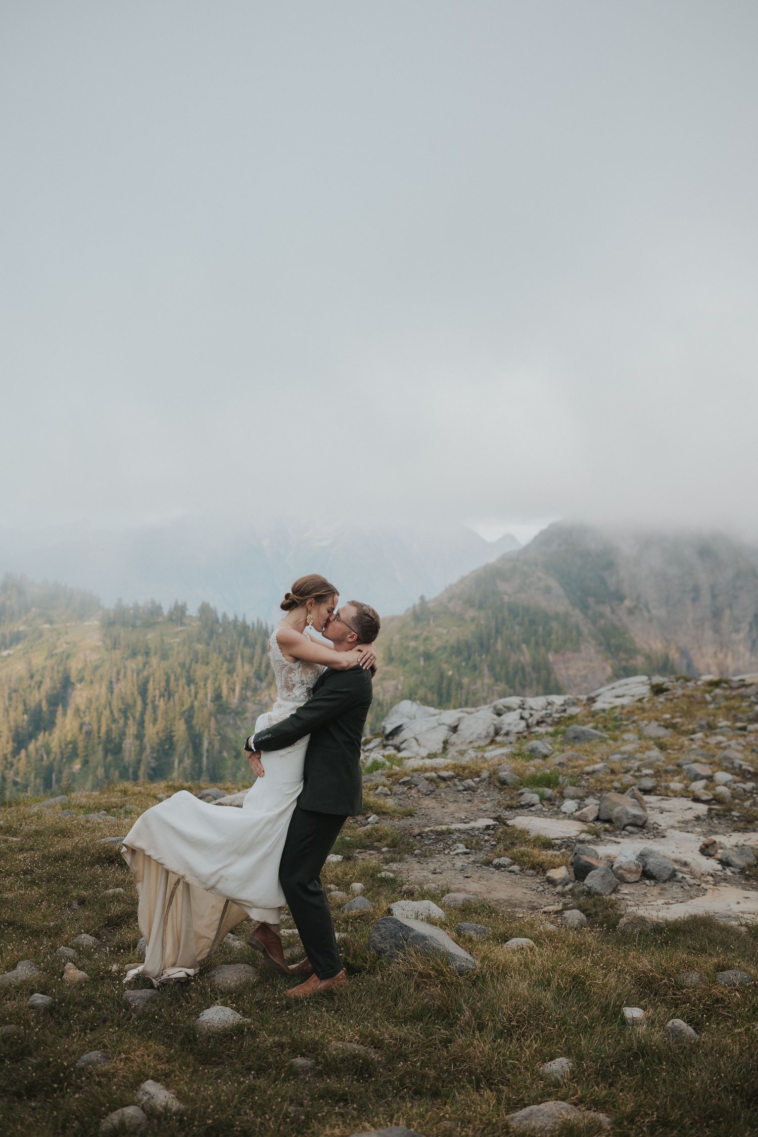 Wedding couple embraces on rocky mountain terrain with dramatic cloudy sky and forest views.