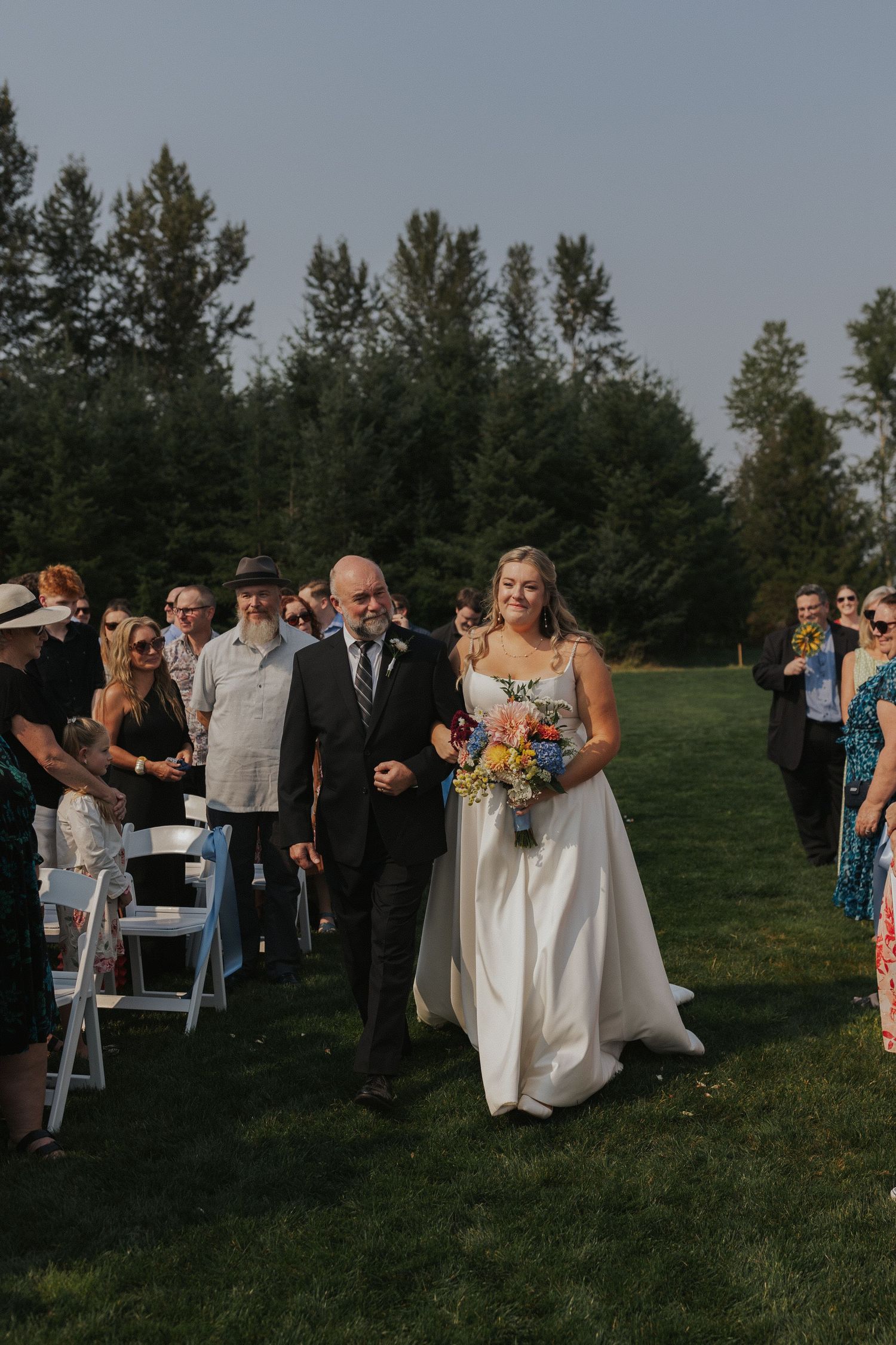 Outdoor wedding ceremony processional on a grassy hill with white chairs and evergreen backdrop.
