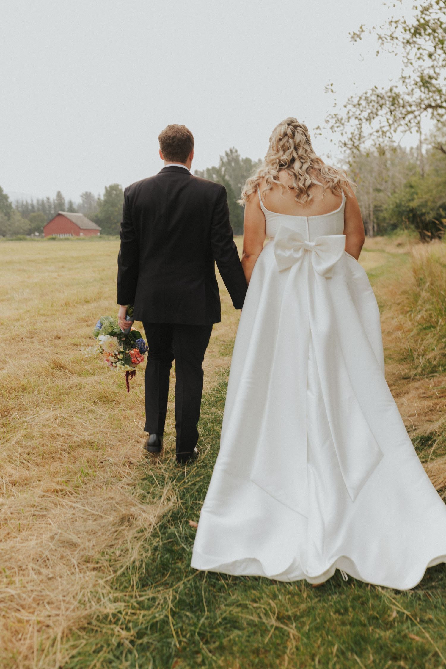 A bride in a flowing white dress and groom walk through a rustic outdoor setting.