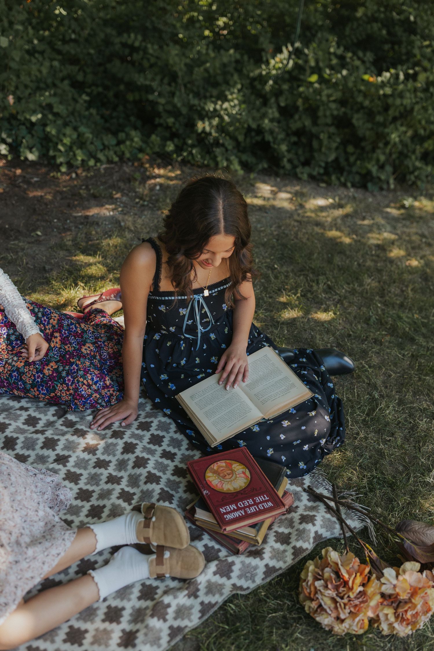 A cozy reading scene with books and dried flowers on a patterned blanket in dappled sunlight.