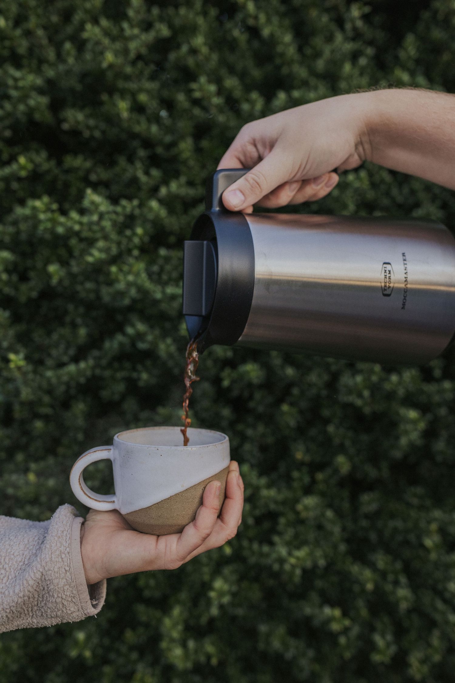 Hands pouring steaming coffee from metal thermos into white ceramic mug against green background.
