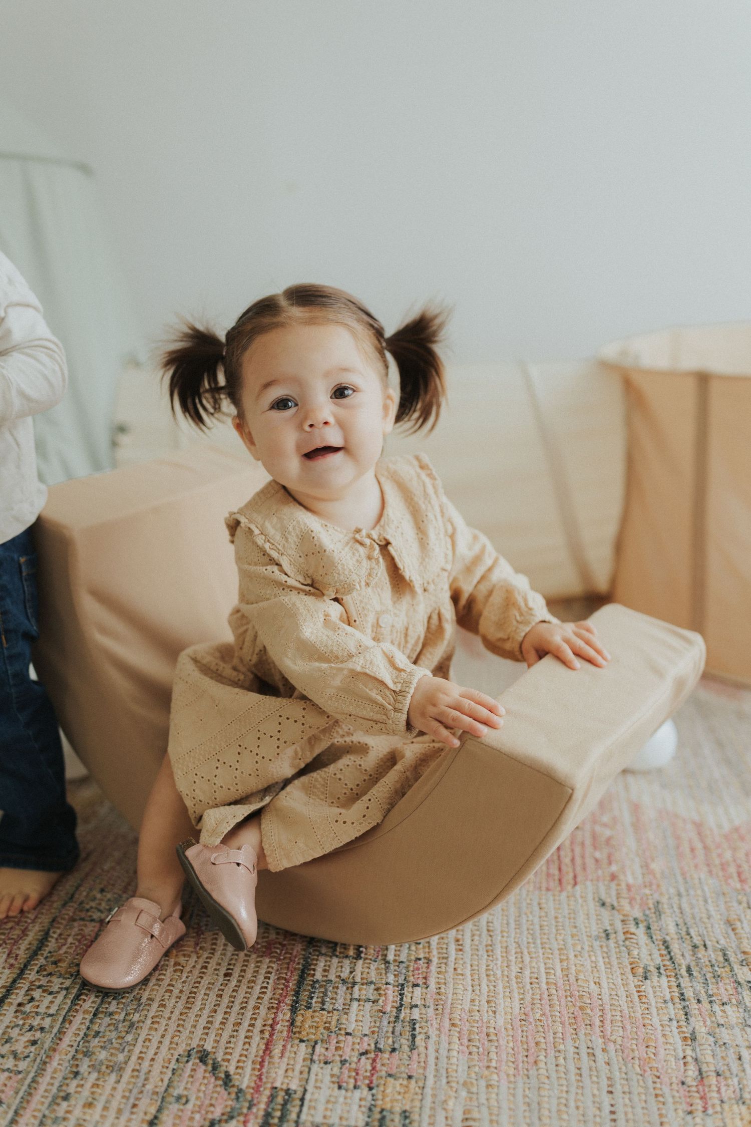 Baby in cream-colored dress sitting on brown paper on patterned carpet.