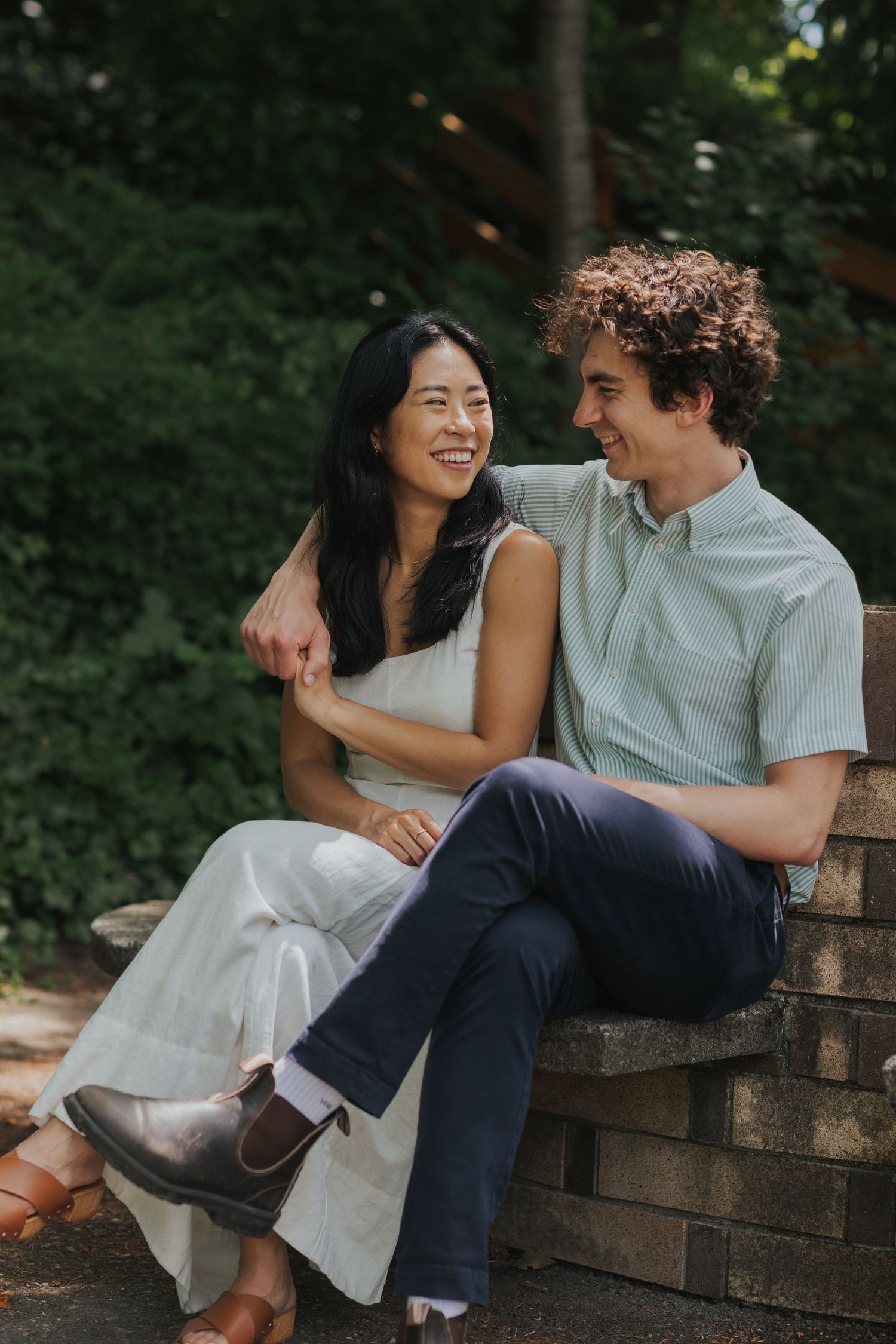 Couple sitting close together on a stone wall surrounded by lush greenery.