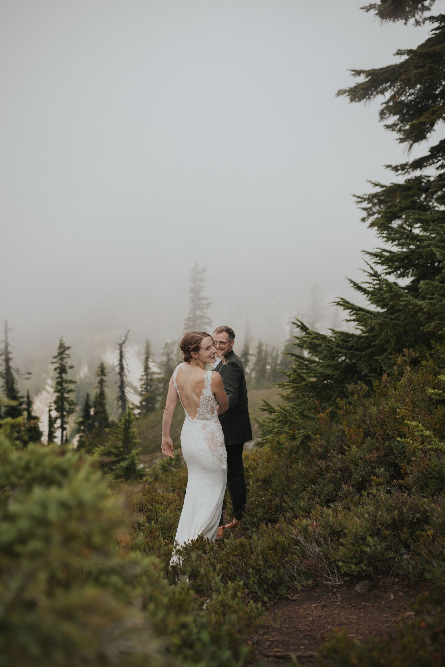 Couple in wedding attire shares intimate moment among evergreen trees in misty mountain setting.