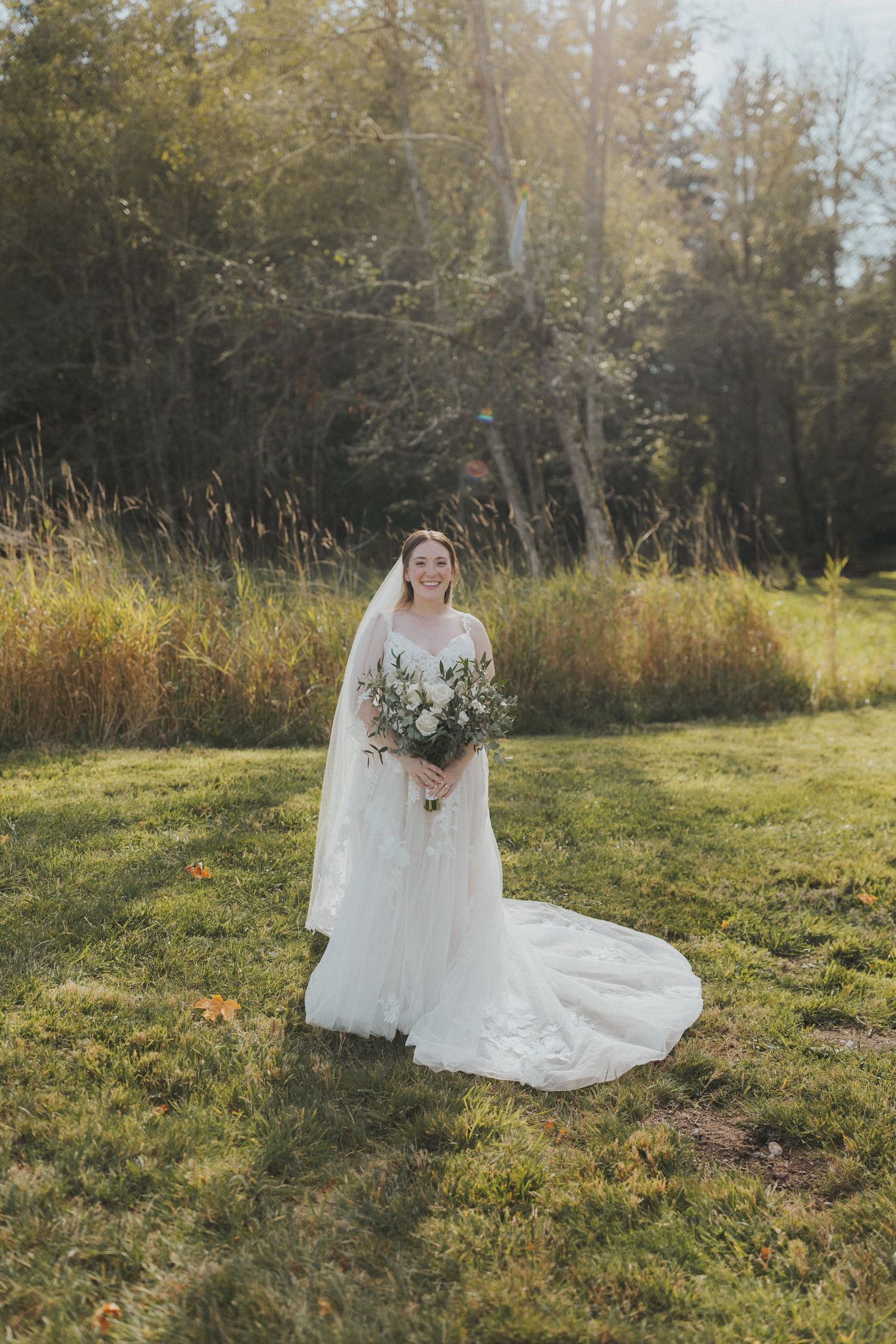 Bride in flowing white dress poses solo in a natural meadow setting during golden hour.