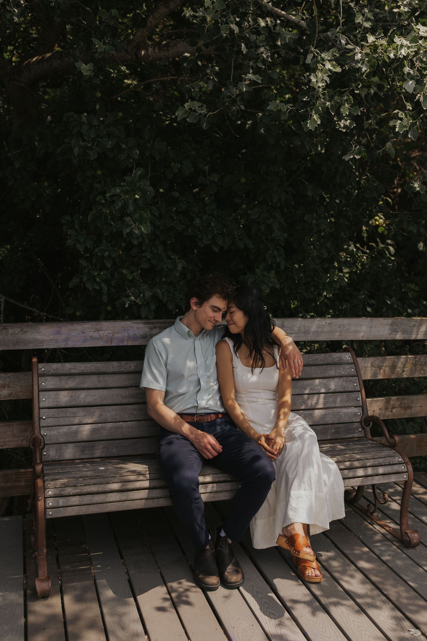 Couple sharing intimate moment on wooden bench in dappled sunlight.