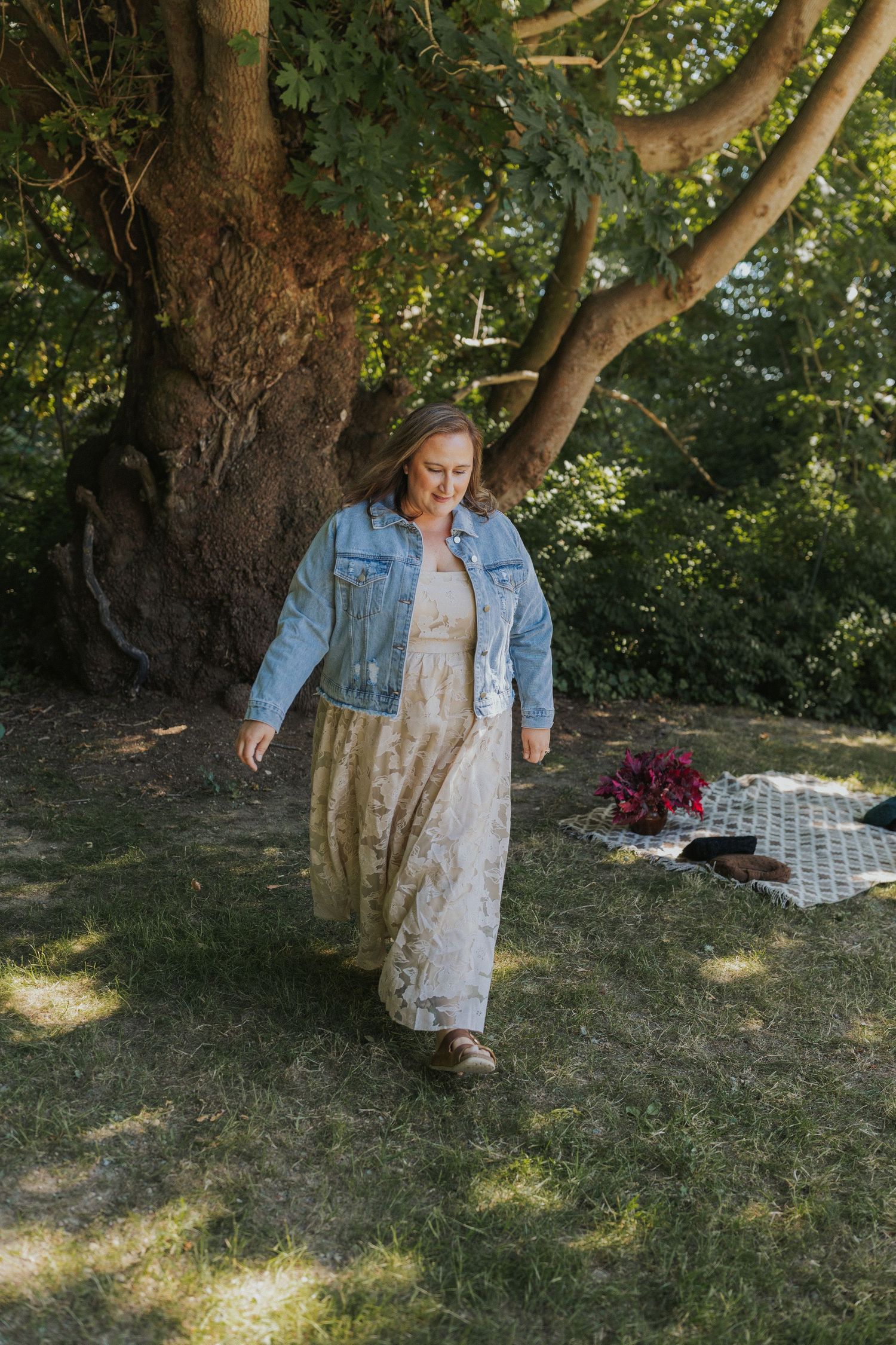 Someone in a denim jacket and vintage-style dress walks across a sunlit grassy area.