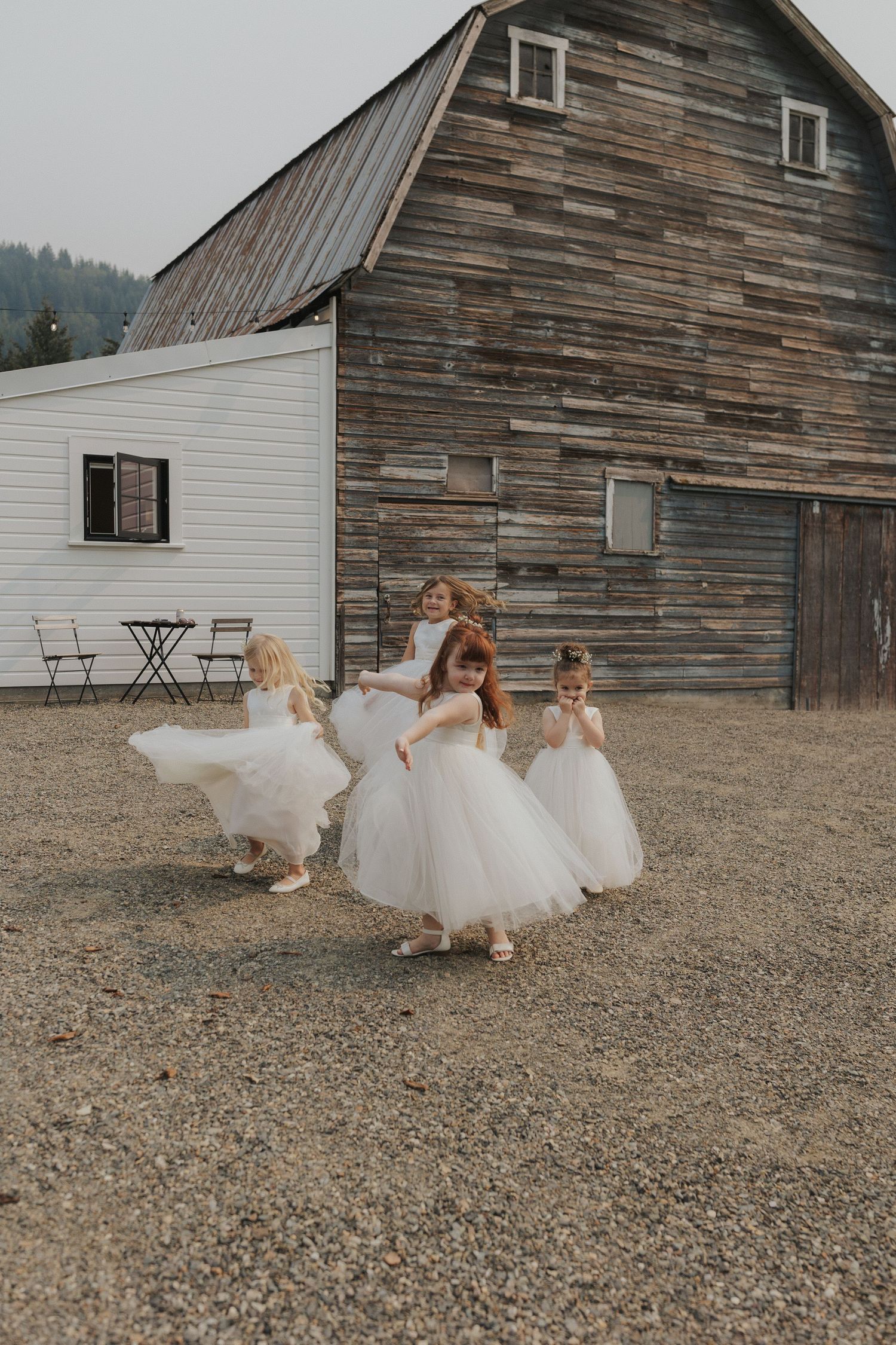 Bridesmaids in white tulle dresses twirl and dance playfully in front of a rustic barn.
